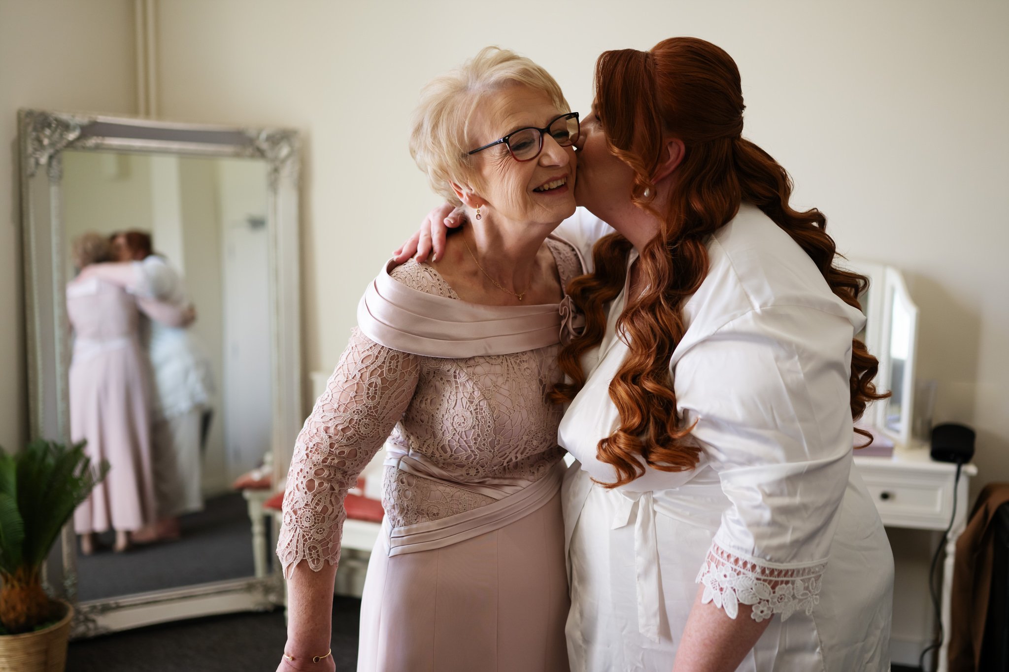 An elderly woman in an elegant dress is being kissed on the cheek by a younger woman with red hair, captured beautifully by the Missenden Abbey wedding photographer. They stand in a room where a mirror reflects another couple in an embrace, adding to the charm of Liz & Olly's wedding at Missenden Abbey.