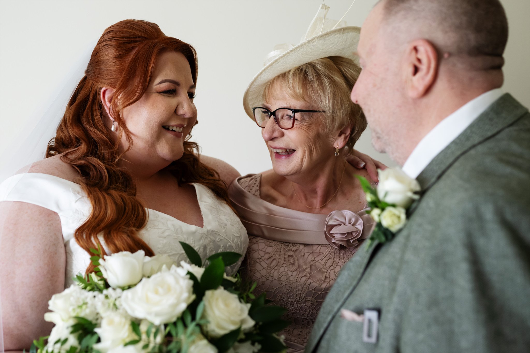 At Liz & Olly's wedding, a bride in a white dress holding a bouquet of white flowers stands happily with an older woman in a beige dress and hat, and an older man in a grey suit.