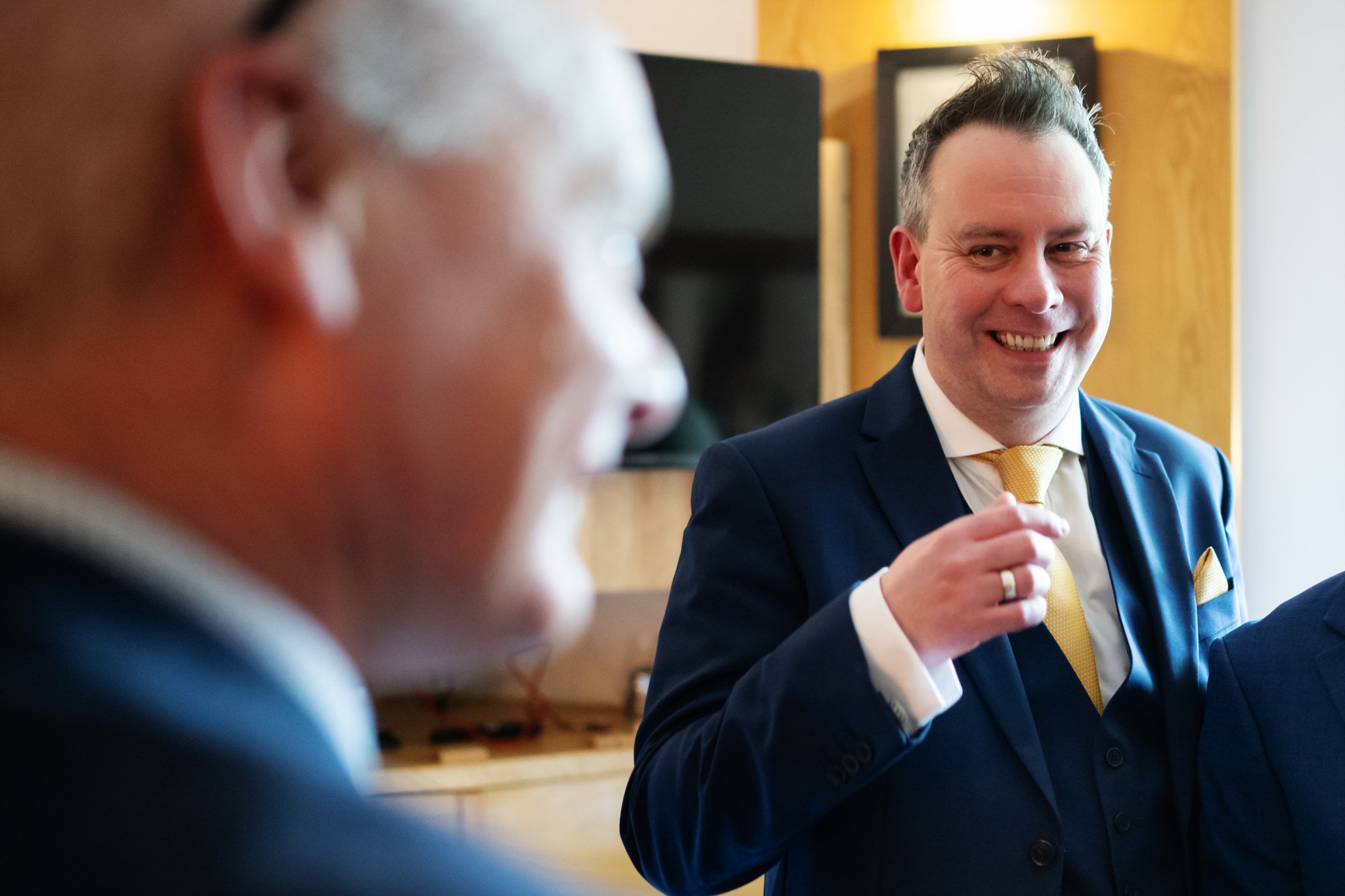 Two men, one in focus and one out of focus, are dressed in formal attire. The man in focus is smiling, wearing a blue suit and a yellow tie, and holding a piece of paper or card at Liz & Olly's wedding at Missenden Abbey.