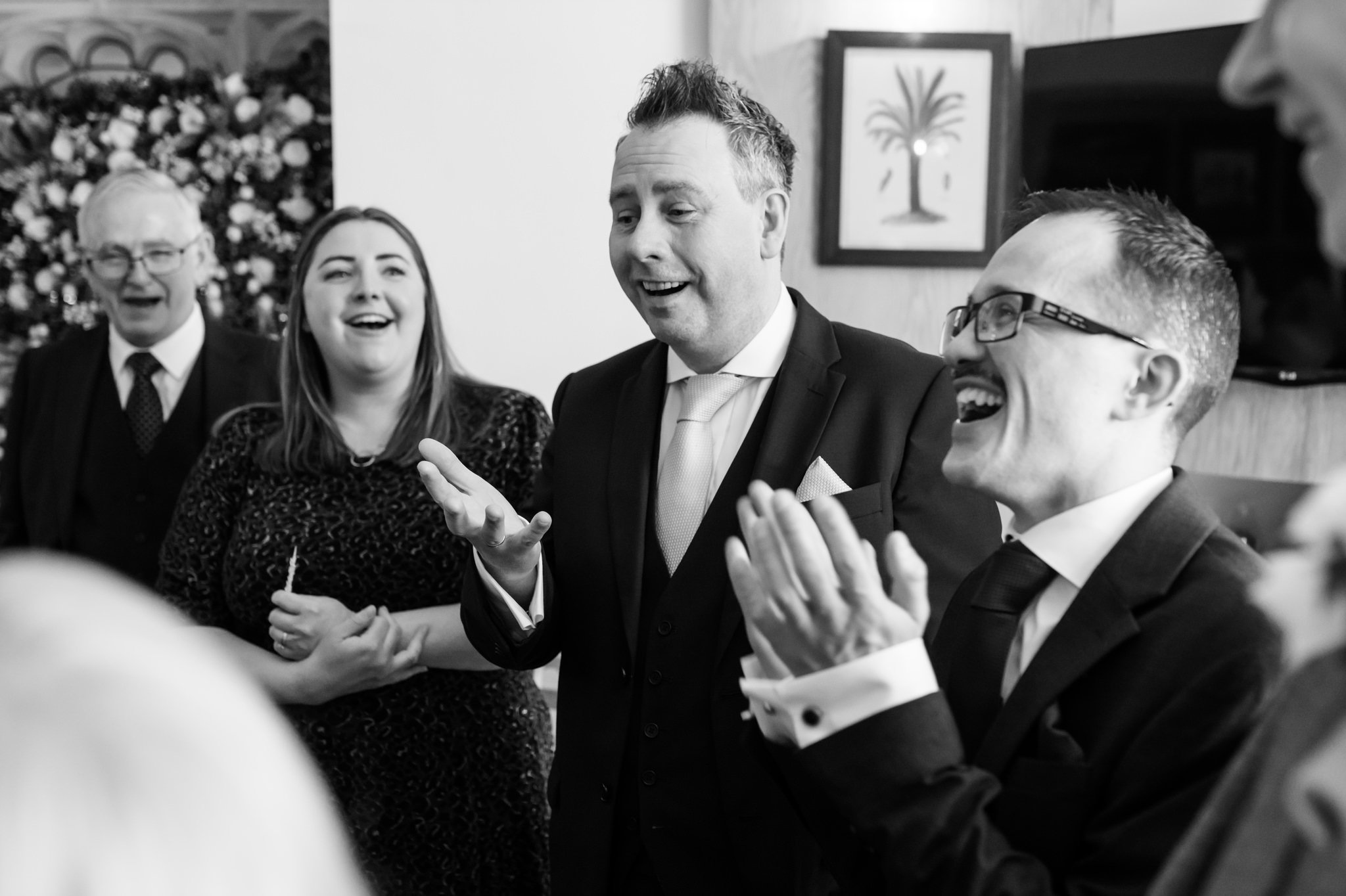 A group of people, dressed formally, are engaged in a lively conversation and laughing at Liz & Olly's wedding. One man gestures with his hands while others smile and clap in this delightful black and white photo.