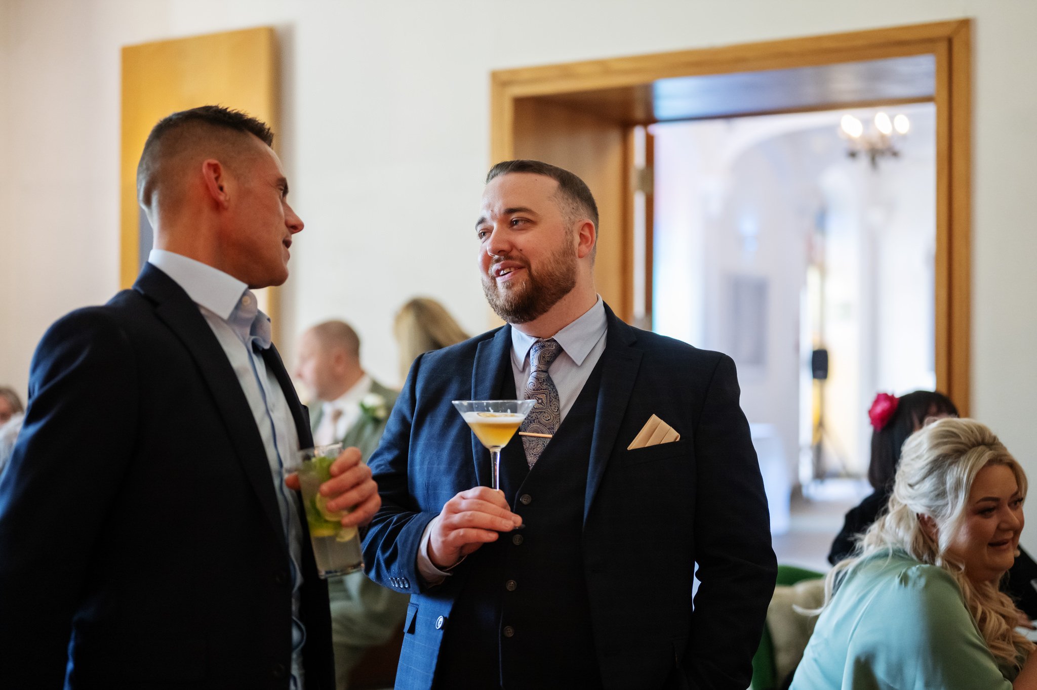 Two men in suits converse while holding cocktails, surrounded by other guests mingling in a casual indoor setting at Liz & Olly's wedding. It's a lively atmosphere captured beautifully by the Missenden Abbey wedding photographer.