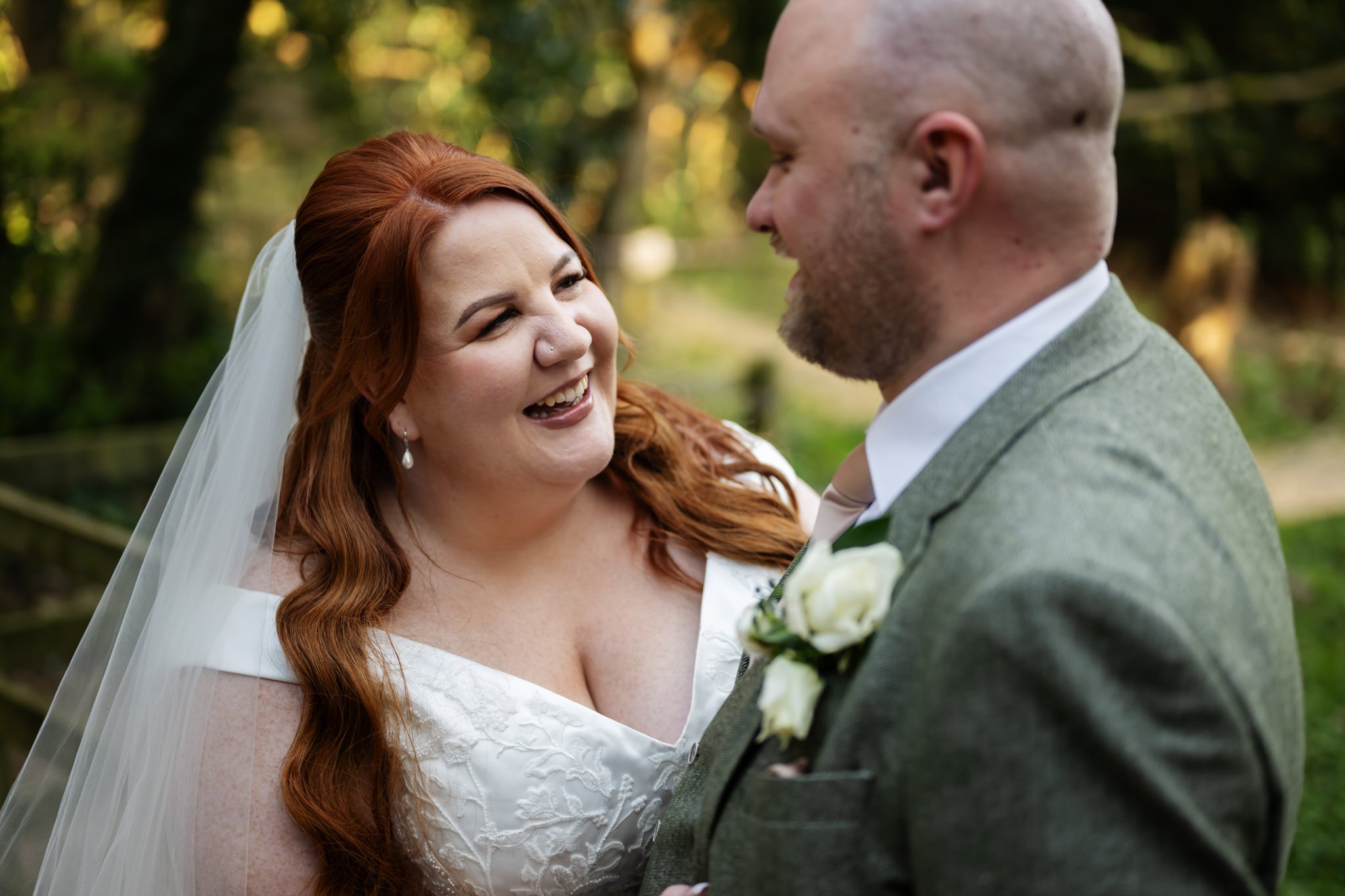 Liz and Olly smile at each other outdoors on their wedding day at Missenden Abbey. The bride, with her red hair flowing beneath a white veil, stands beside her groom in a gray suit adorned with a white boutonniere. Their joy is beautifully captured by the Missenden Abbey wedding photographer.