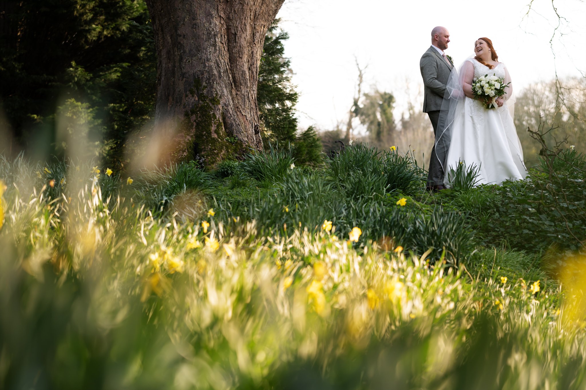 A bride and groom stand beside a large tree in a garden setting at Missenden Abbey, surrounded by greenery and yellow flowers. The bride holds a bouquet, and both are looking at each other with adoration. This beautiful snapshot captures the magic of Liz & Olly's wedding day.