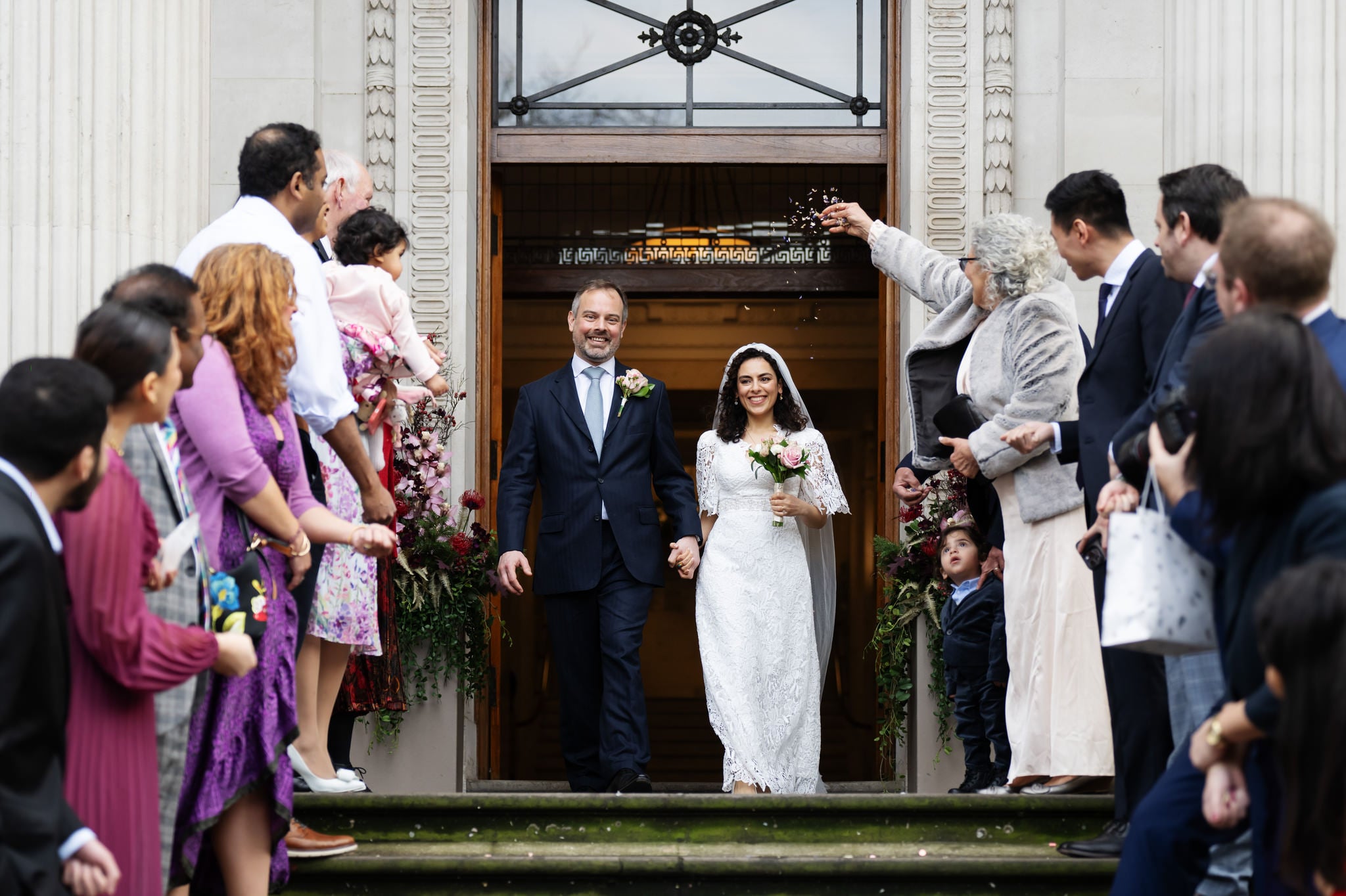 A newlywed couple, Dana & Phil, exits the Old Marylebone Town Hall while guests cheer and throw confetti. The bride wears a white dress and the groom a blue suit. They are surrounded by friends and family on both sides, capturing every joyous moment with their wedding photographer.