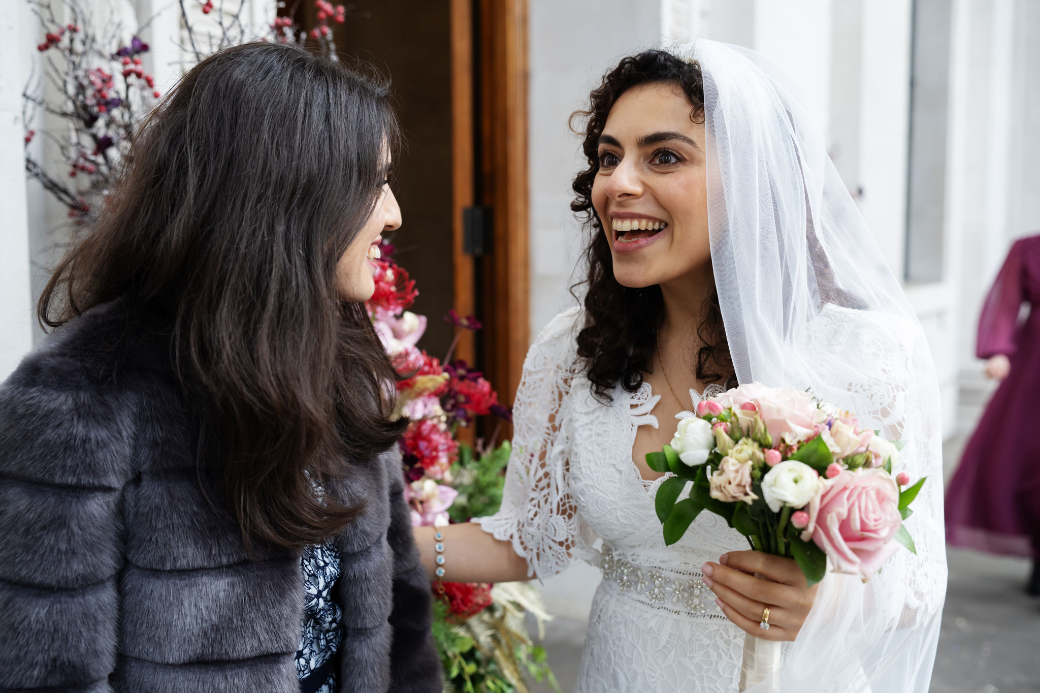 Two women are talking and smiling in front of a building. One woman is dressed in a wedding gown and holding a bouquet, while the other is wearing a dark fur coat. They are celebrating an Old Marylebone Town Hall Wedding, beautifully captured by their talented wedding photographer.