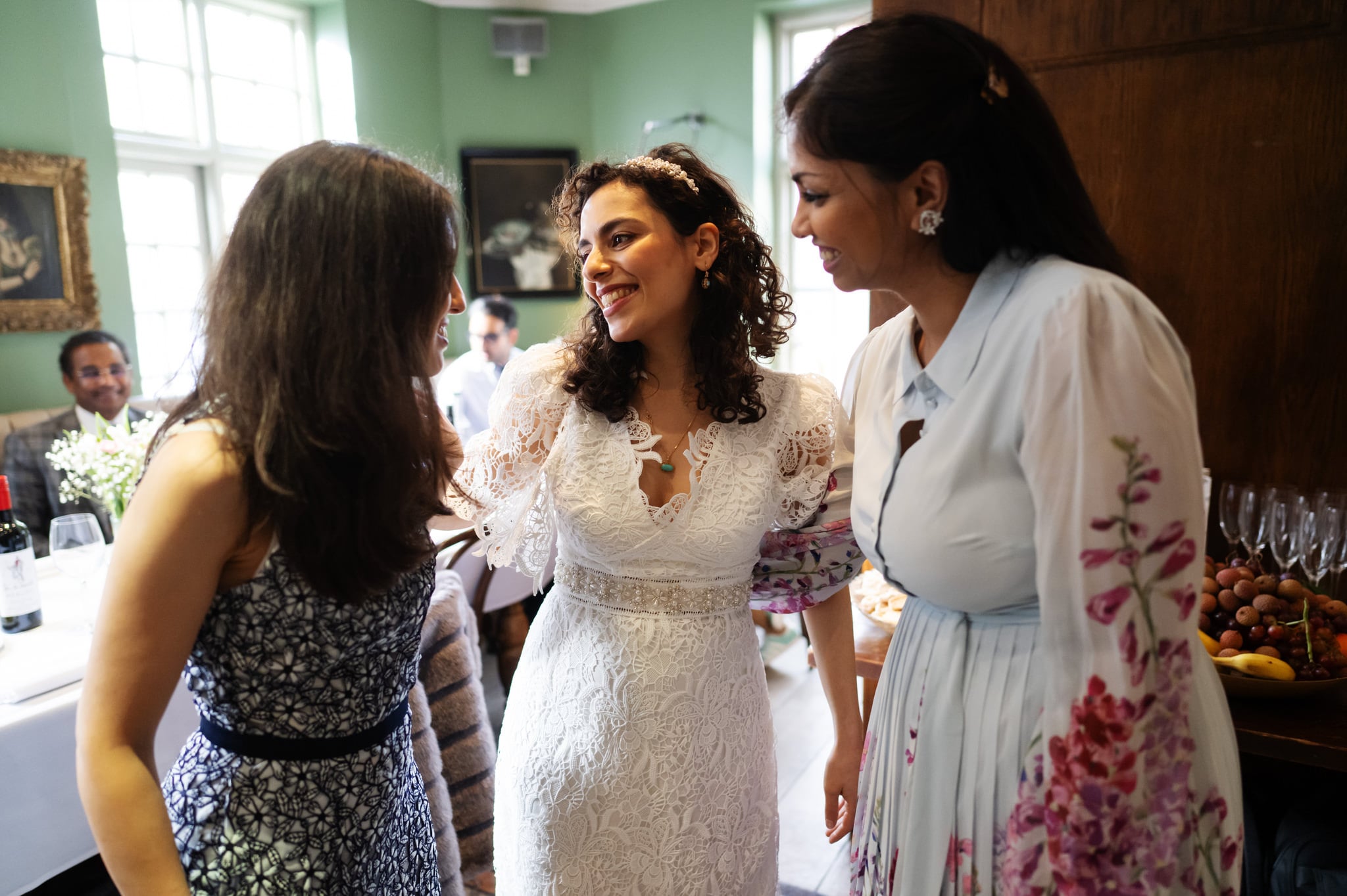 Three women are conversing in a well-lit room. The woman in the center, wearing a white lace dress, is smiling. The others are wearing patterned dresses. A table with glasses and fruit is in the background, perfectly captured by the wedding photographer at Dana & Phil's wedding in London.