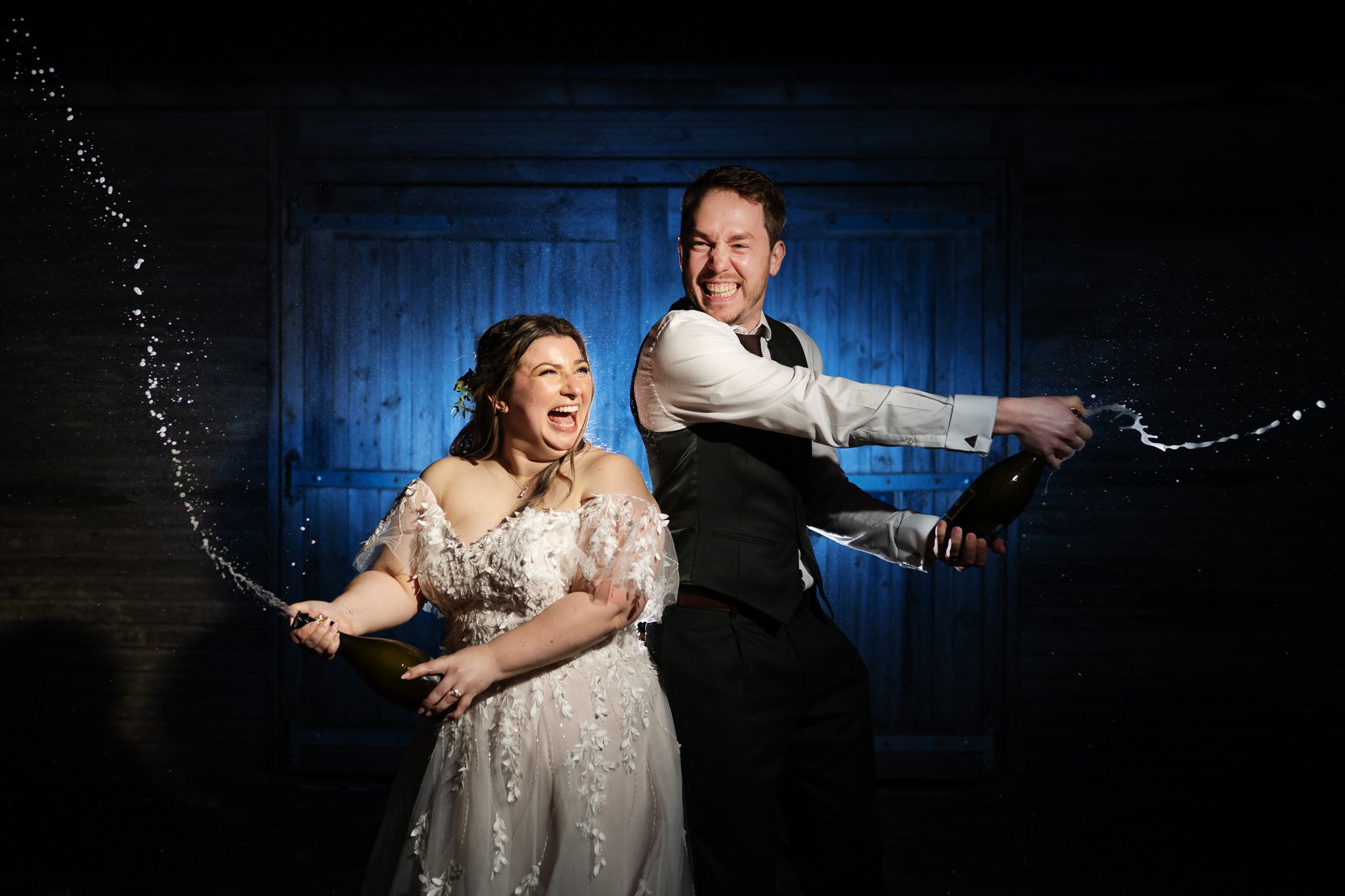 A bride and groom joyfully spray champagne from bottles. They are standing in front of a wooden backdrop, at Stratton Court Barn. The couple are smiling and laughing.