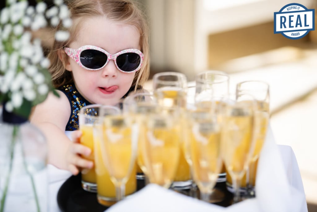 A child wearing sunglasses reaches for a glass of orange juice from a tray filled with glasses. The girl is sticking her tongue out in anticipation. Captured at St Michaels Manor.