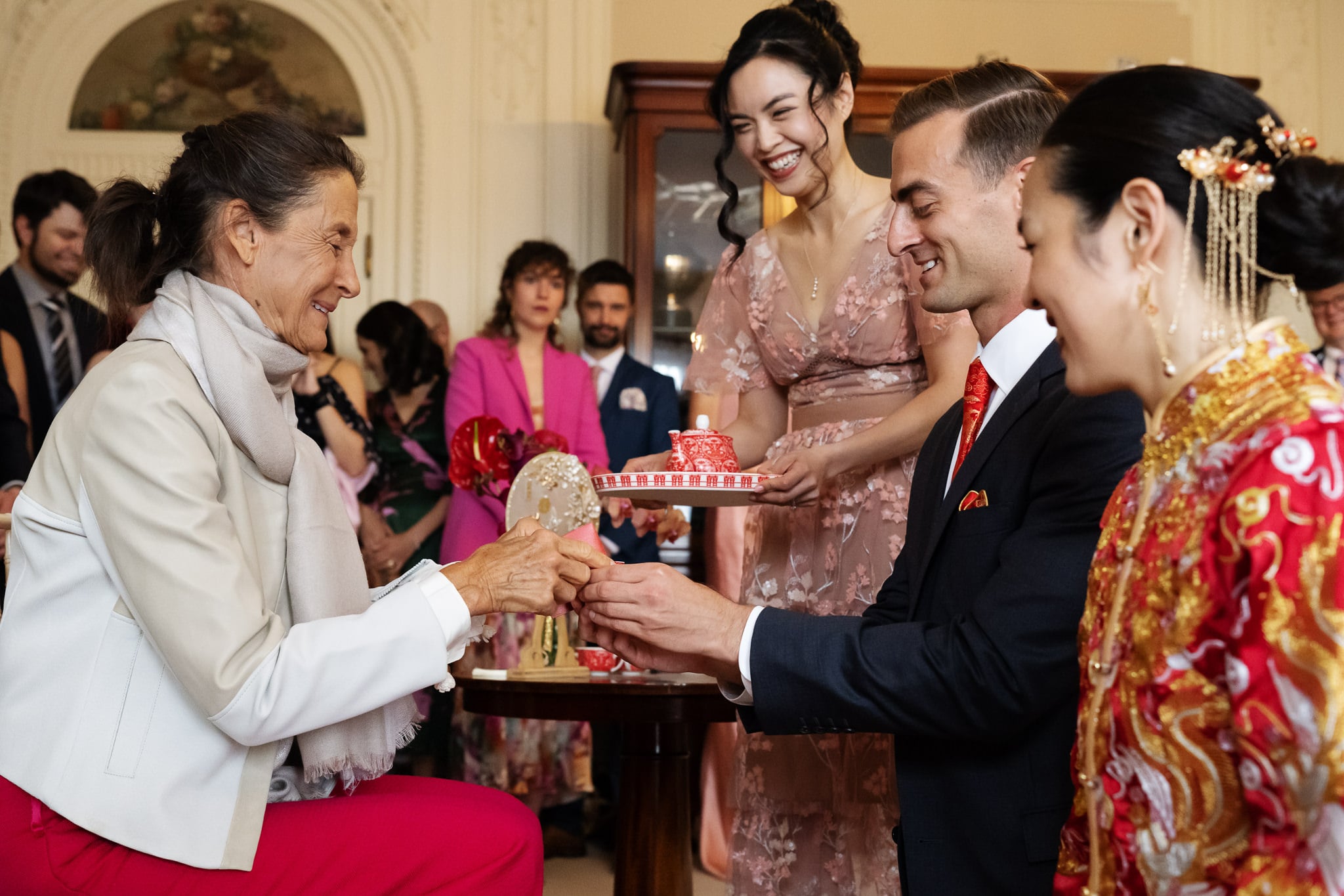A couple in formal attire kneel and present tea to a seated woman during a traditional tea ceremony at No. 4 Hamilton Place, with London wedding guests watching in the background.