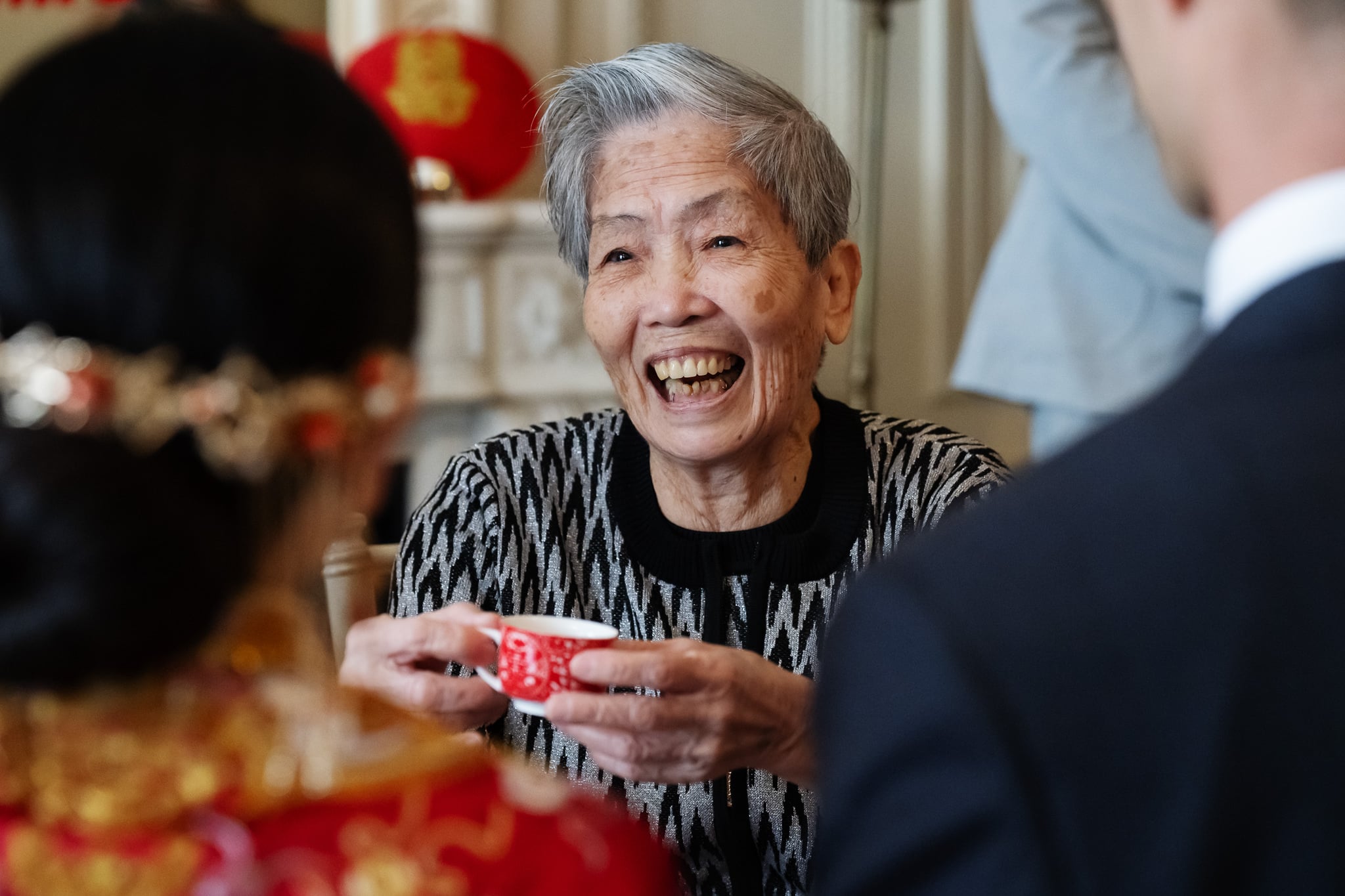 An elderly woman with gray hair smiles while holding a red and white teacup at No. 4 Hamilton Place, facing two people dressed in formal attire during a London wedding.