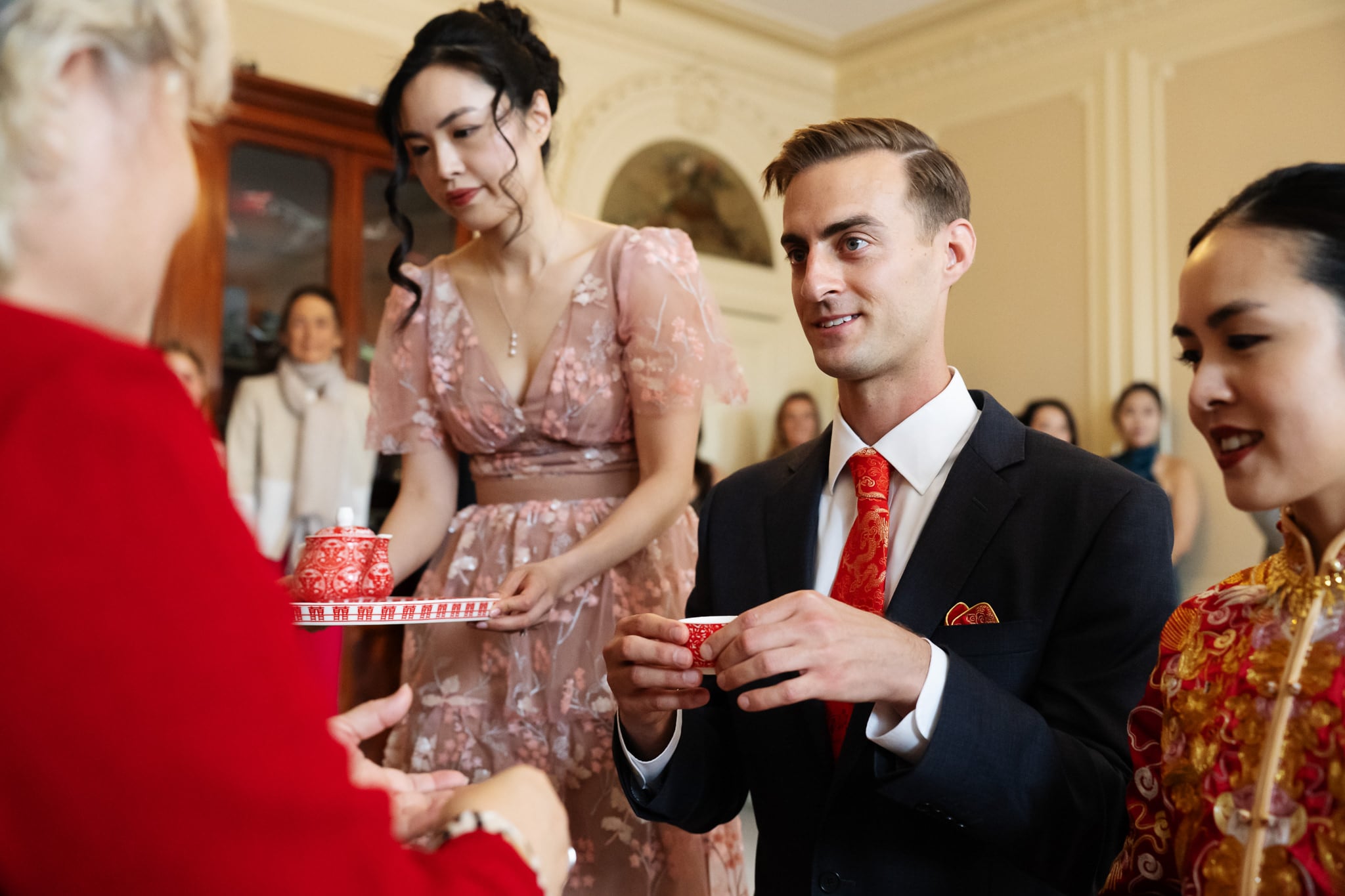 A man in a suit and two women in traditional attire participate in a tea ceremony, holding red teacups in a formal indoor setting at No. 4 Hamilton Place, perfect for a refined London wedding.