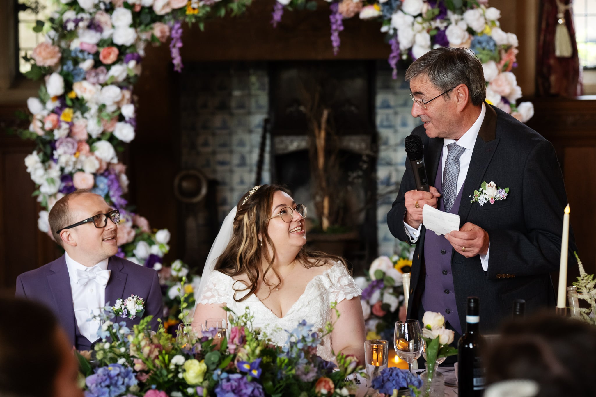 At Fanhams Hall, a man in a suit gives a speech with a microphone at the wedding reception while the bride and groom, seated at a flower-adorned table, smile and listen.