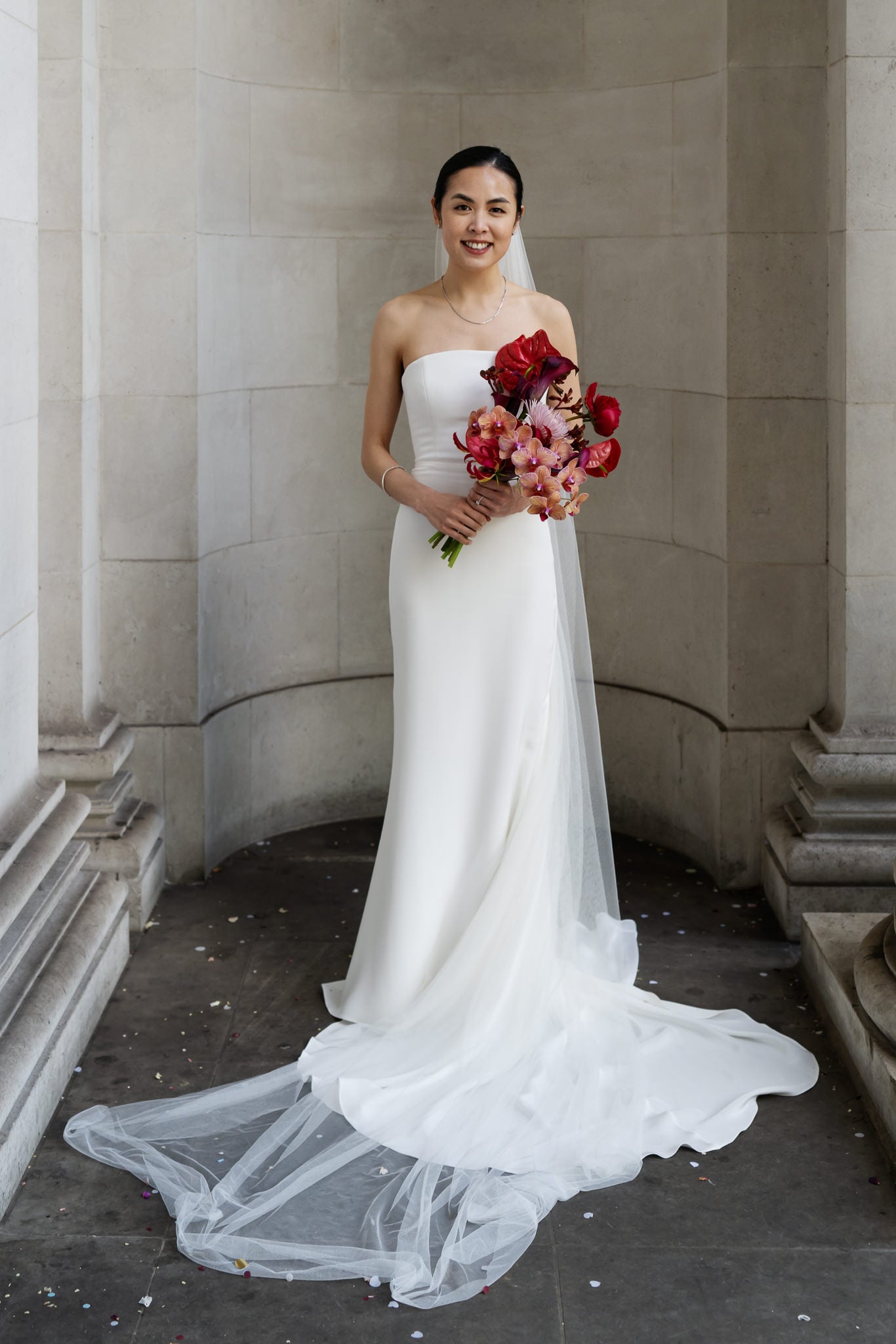 A woman in a strapless white wedding dress holds a bouquet of red and pink flowers at No. 4 Hamilton Place, her long veil spread on the ground—capturing the elegance of a London wedding against a stone wall backdrop.
