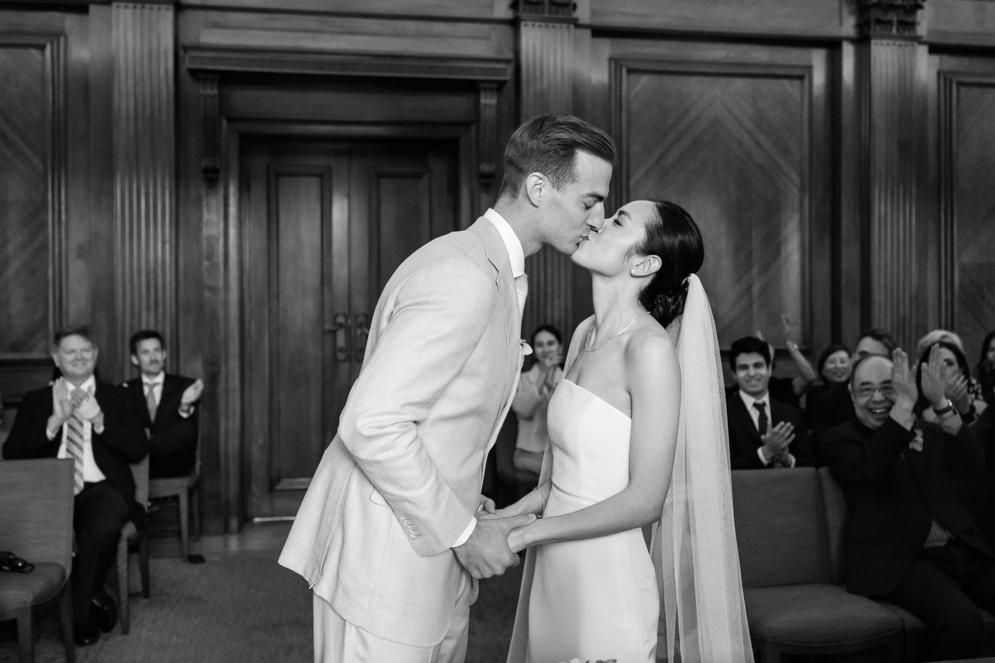 A bride and groom kiss at the altar during a London wedding at No. 4 Hamilton Place, while seated guests applaud in the background at this formal indoor ceremony.