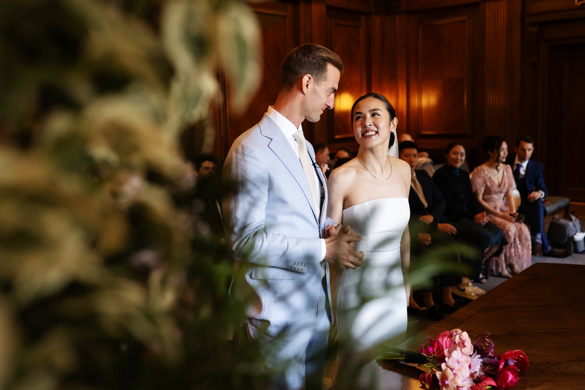 A bride and groom stand together smiling in a wood-paneled room at No. 4 Hamilton Place, with guests seated in the background and a bouquet on the table, capturing an elegant London wedding moment.