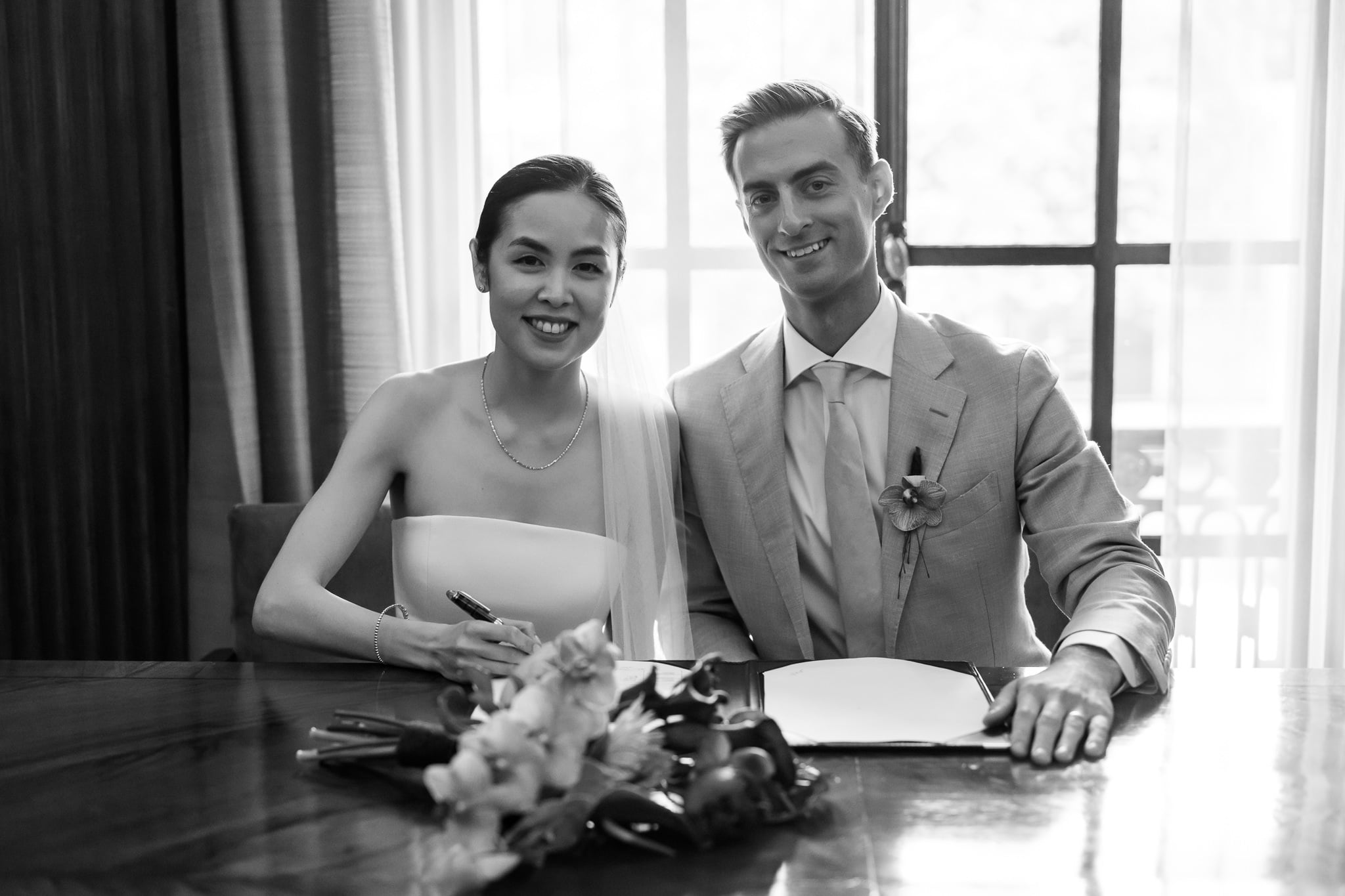 A couple sits at a table at No. 4 Hamilton Place, smiling at the camera with a document and bouquet in front of them, ready to celebrate their London wedding. The woman holds a pen, and both are dressed formally.