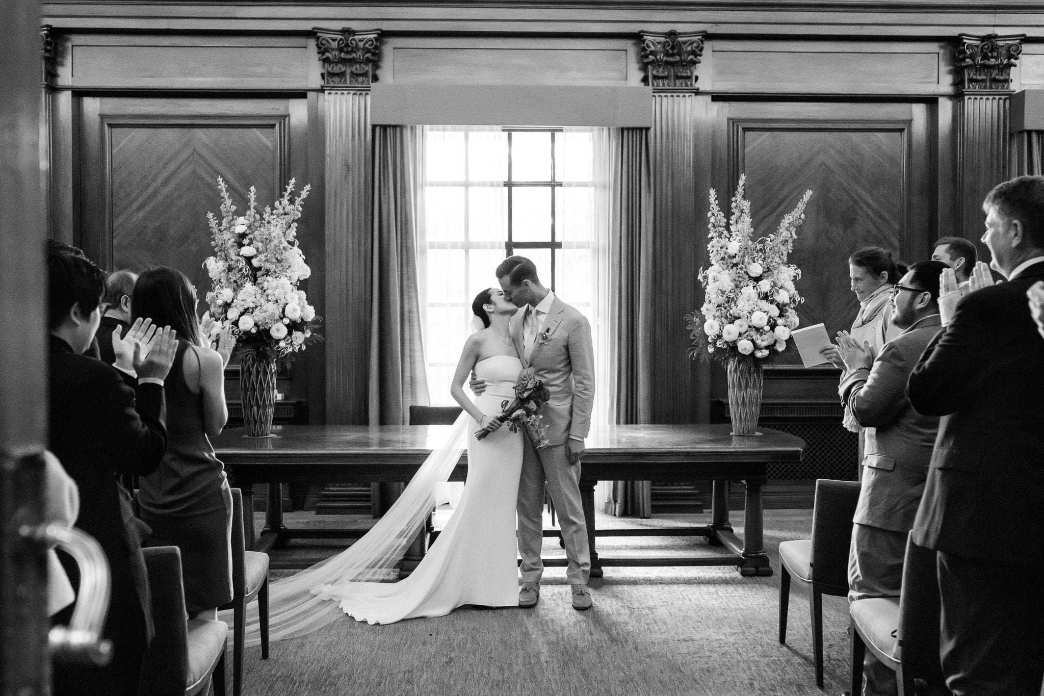 A bride and groom kiss at the altar during a formal indoor London wedding at No. 4 Hamilton Place, surrounded by guests applauding and taking photos.