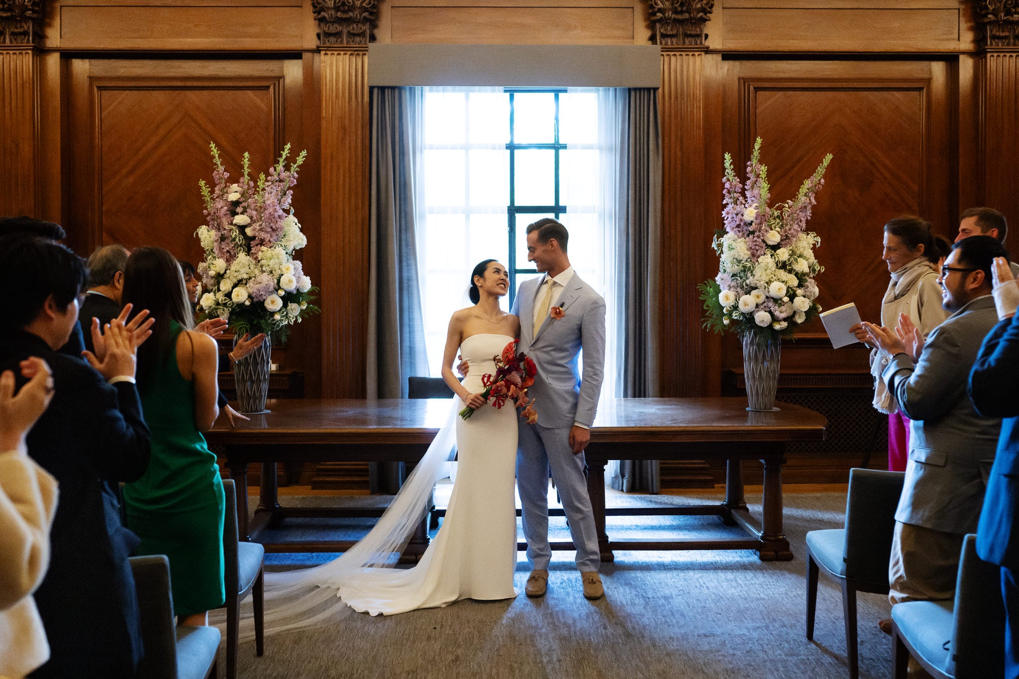 A bride and groom stand together at the front of a wood-paneled room at No. 4 Hamilton Place, surrounded by floral arrangements, as guests applaud their London wedding ceremony.