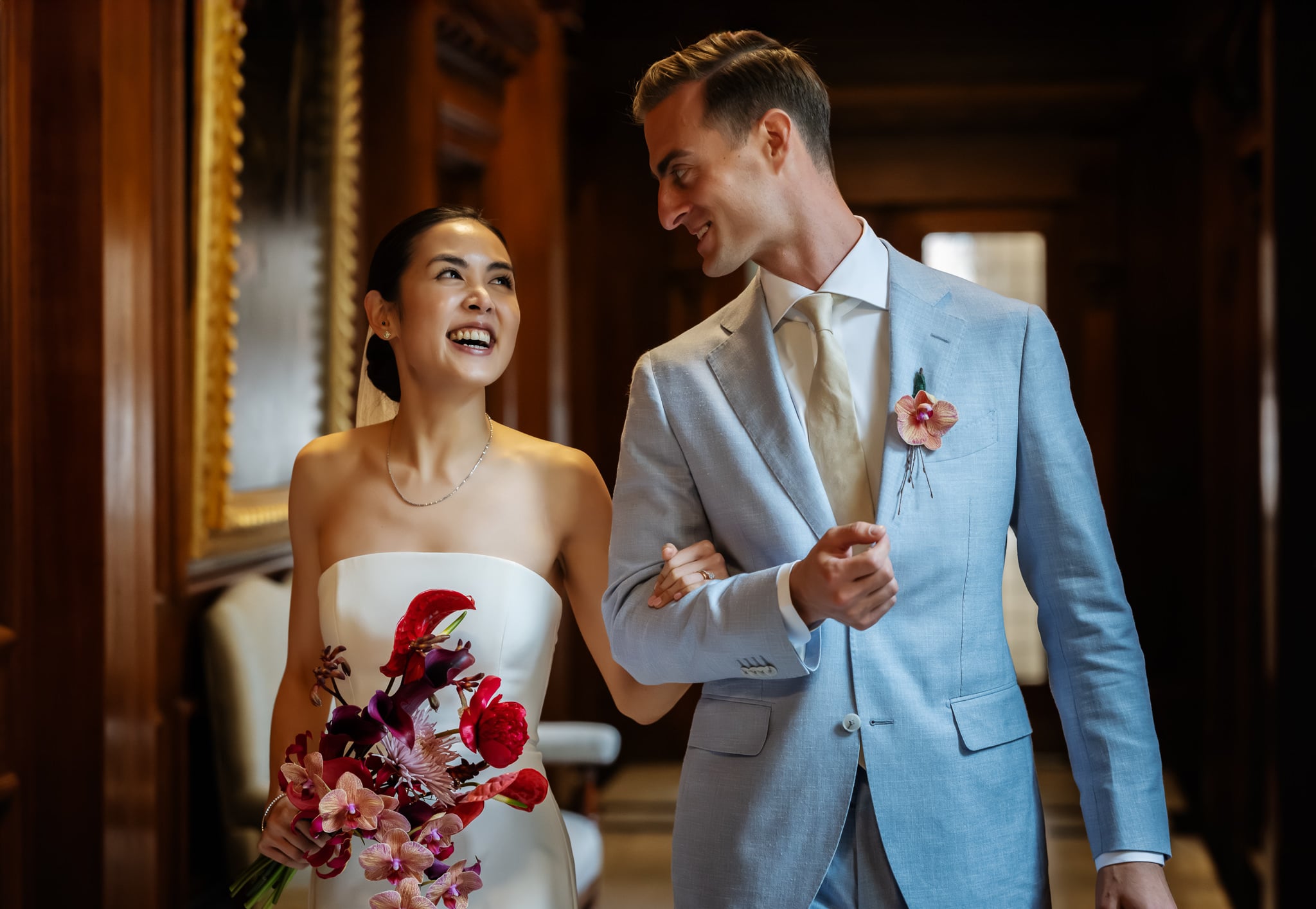 A bride in a white dress holding a bouquet and a groom in a light blue suit walk arm in arm indoors at No. 4 Hamilton Place, both smiling and looking at each other during their beautiful London wedding.