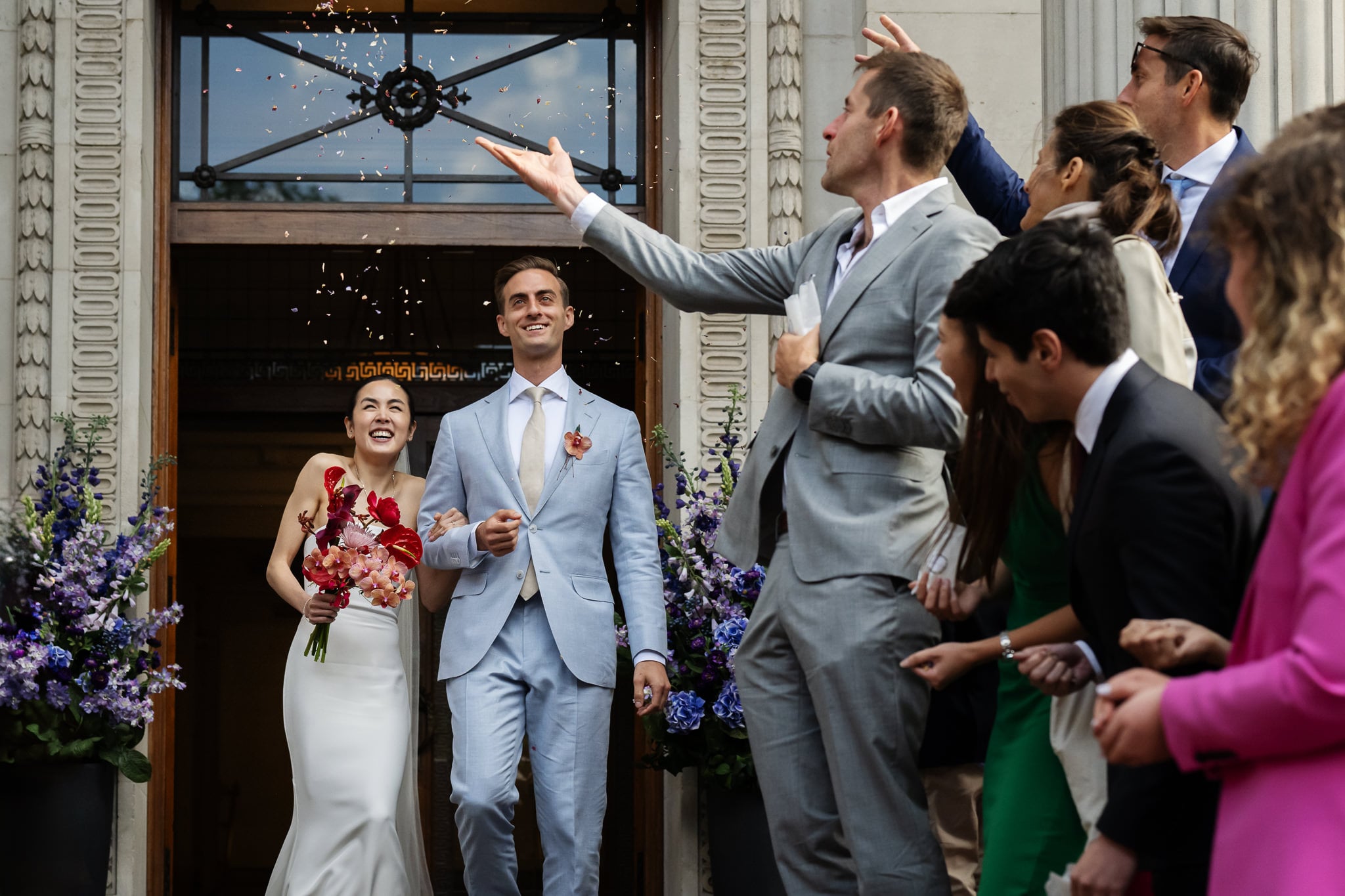 A bride and groom exit No. 4 Hamilton Place at their London wedding as guests in formal attire toss confetti. The couple is smiling, and the entrance is adorned with purple flower arrangements.