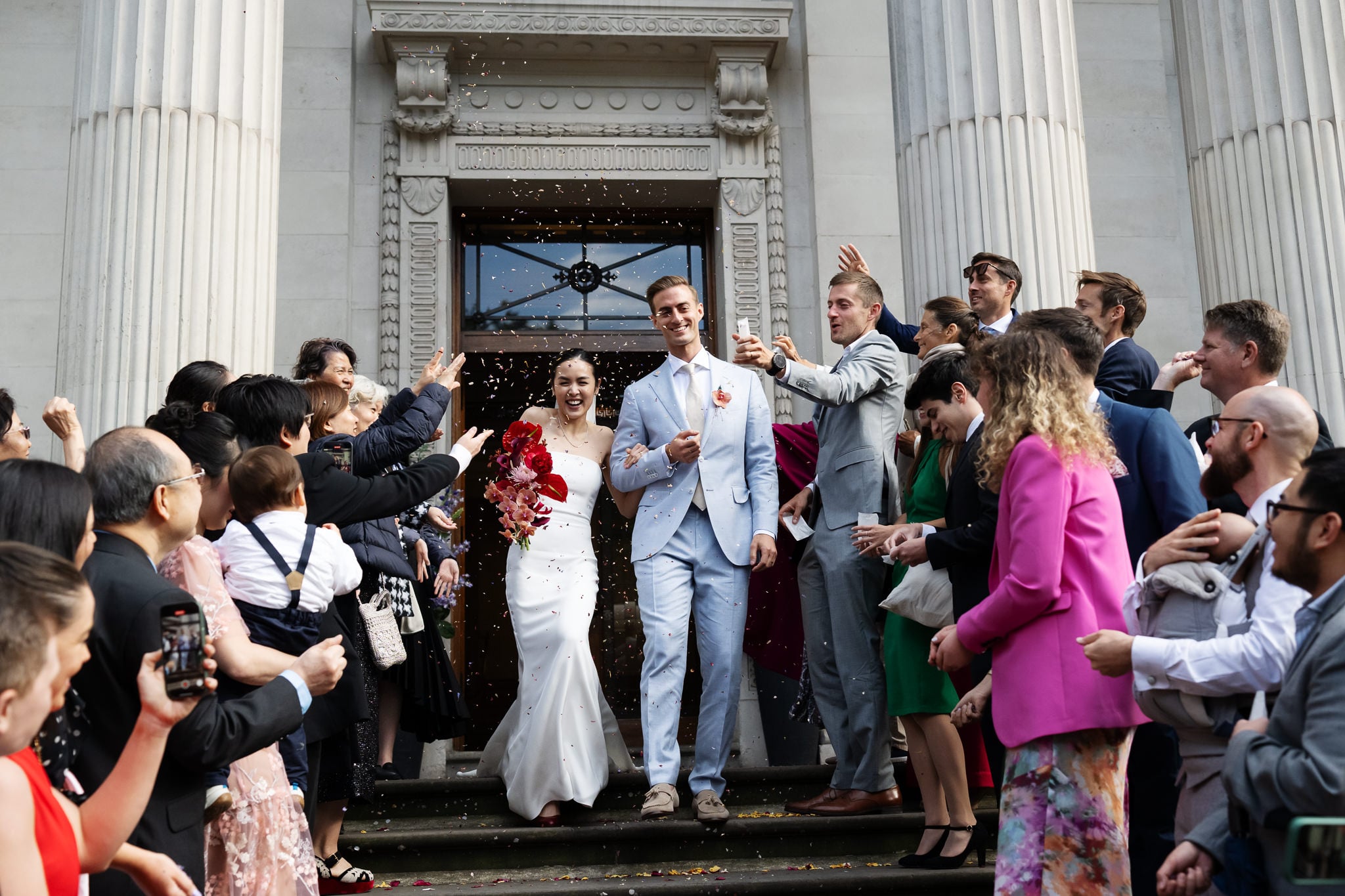 A bride and groom in formal attire exit No. 4 Hamilton Place as guests throw confetti and cheer, standing on steps between two large columns after their London wedding.