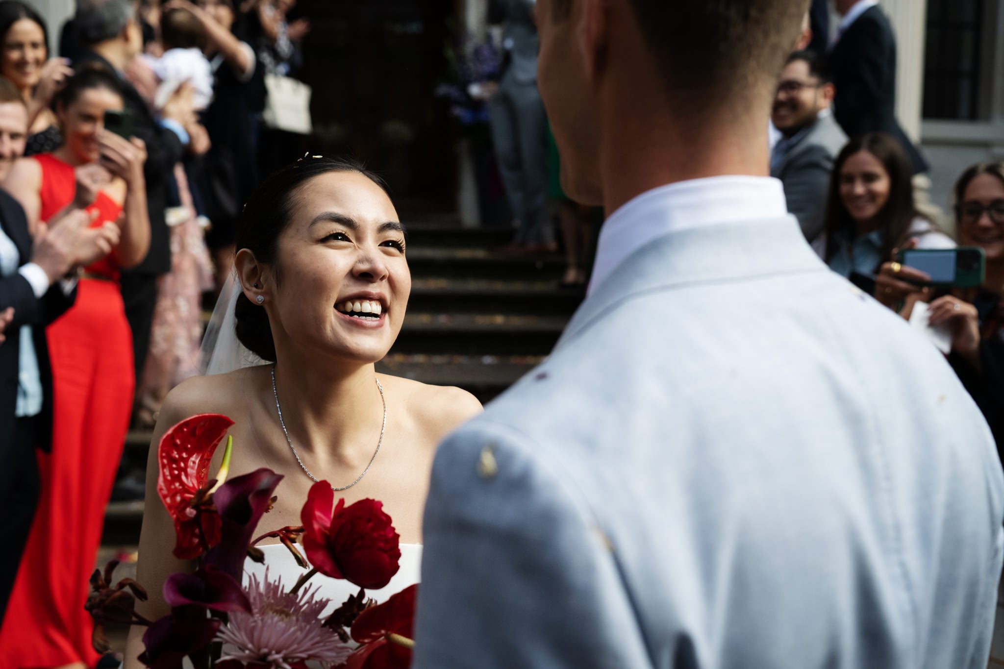 A bride smiles at her partner while holding a bouquet, surrounded by guests applauding outside No. 4 Hamilton Place at a beautiful London wedding.