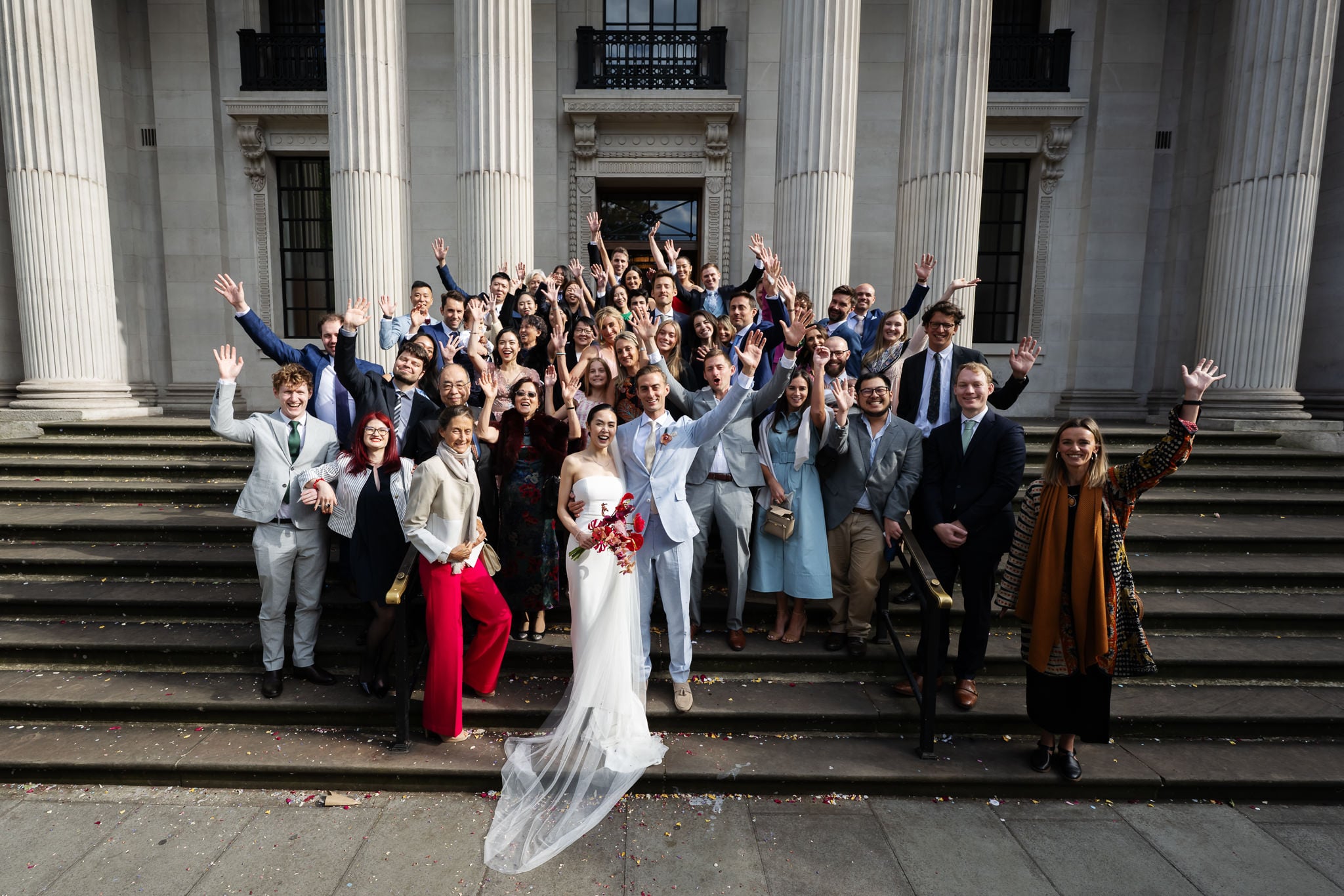 A large group of people, including a bride and groom, stand on steps outside No. 4 Hamilton Place, posing and waving for a group photo at their London wedding.