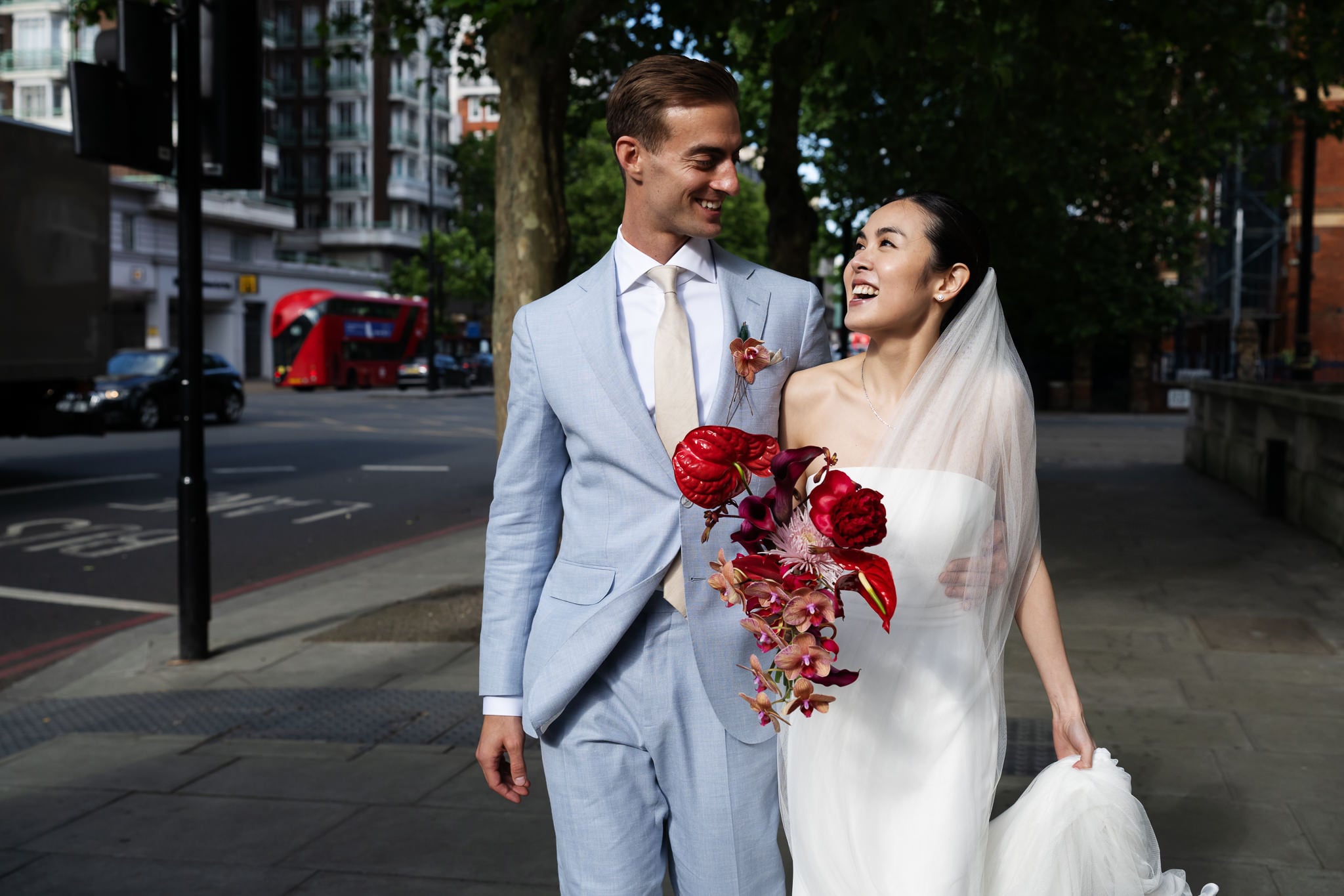 A bride and groom walk outdoors in formal wedding attire, smiling at each other. The bride holds a bouquet of red and pink flowers. Urban street scene with trees in the background, capturing a classic London wedding near No. 4 Hamilton Place.