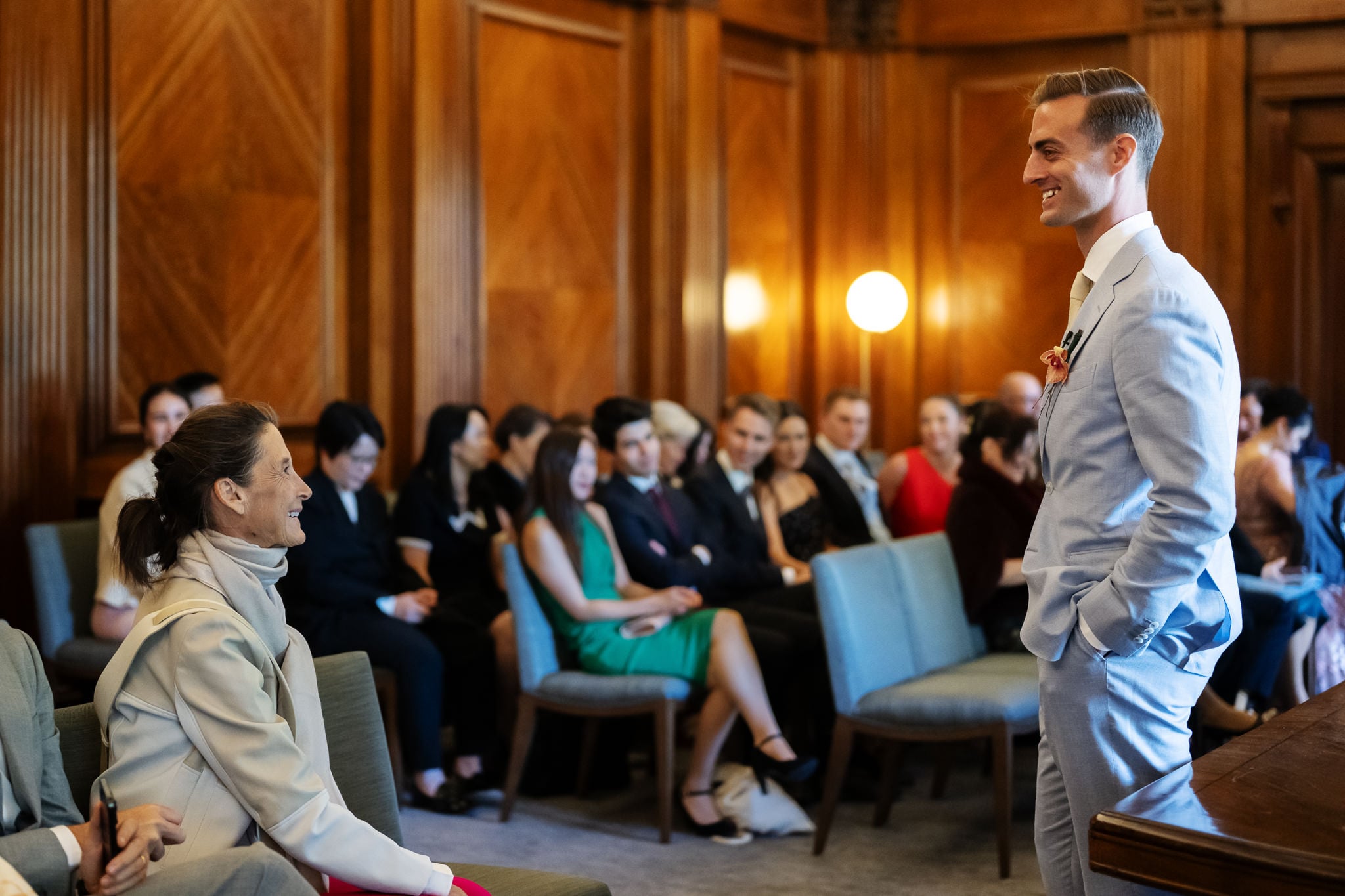 A man in a light suit stands smiling at a seated woman in a wood-paneled room at No. 4 Hamilton Place, with an audience watching and smiling in the background, capturing a joyful London wedding moment.