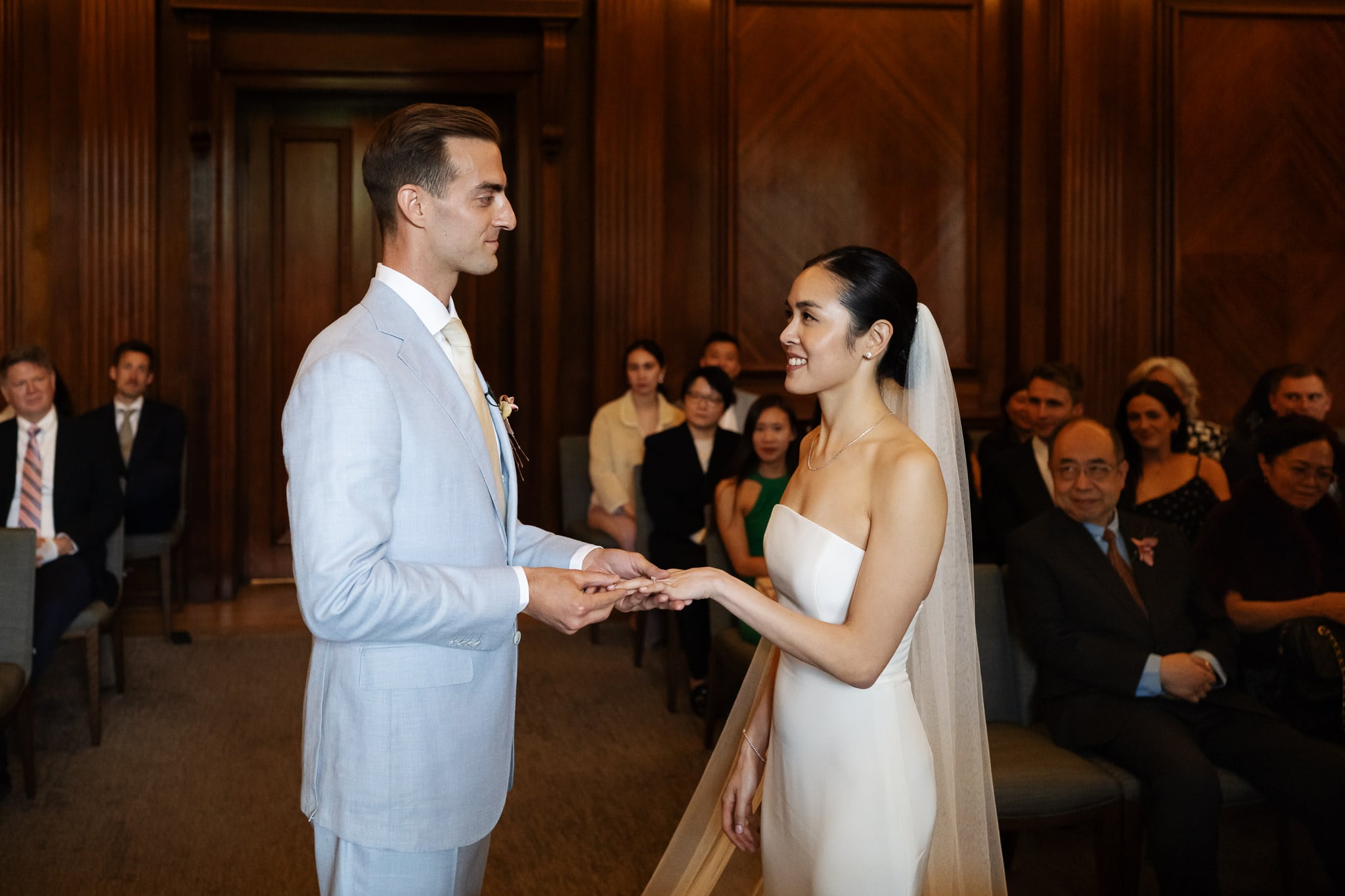 A bride and groom stand facing each other, holding hands, during a London wedding ceremony at No. 4 Hamilton Place in a wood-paneled room with seated guests watching.