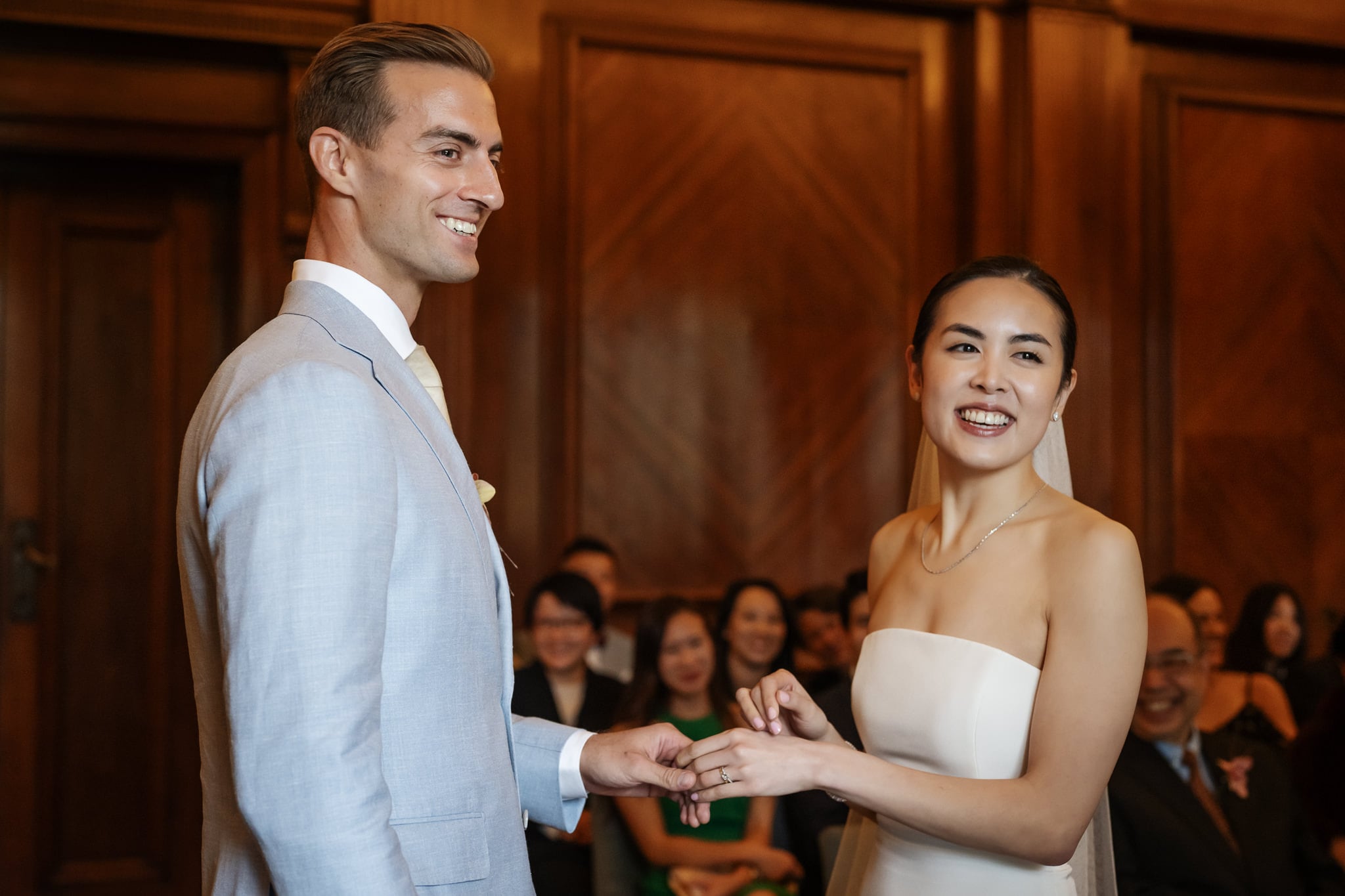 A bride and groom exchange rings during their London wedding at No. 4 Hamilton Place in a wood-paneled room, smiling as guests watch in the background.