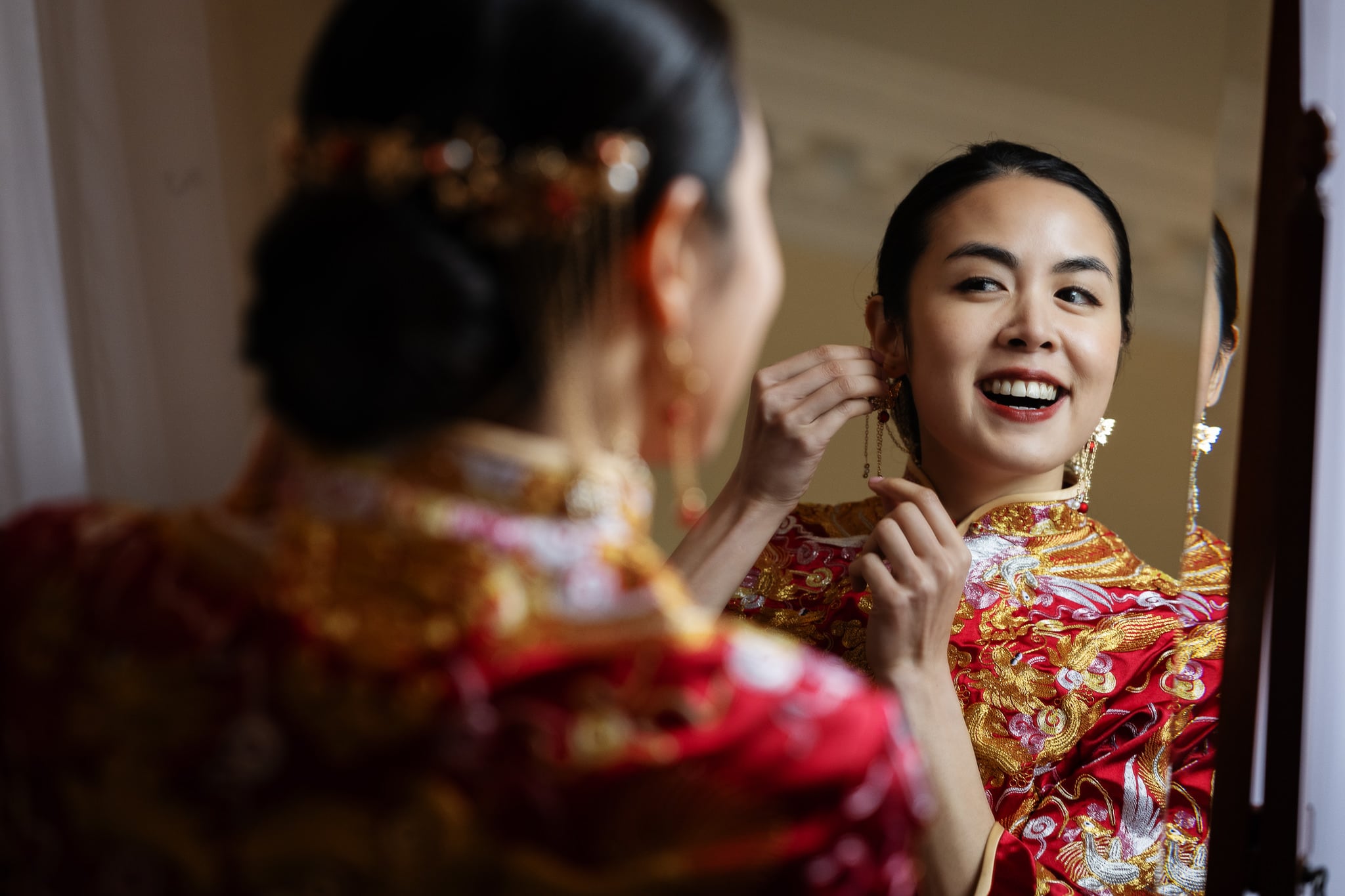 A woman in a red and gold traditional outfit smiles as she adjusts an earring while looking at herself in a mirror before her London wedding at No. 4 Hamilton Place.