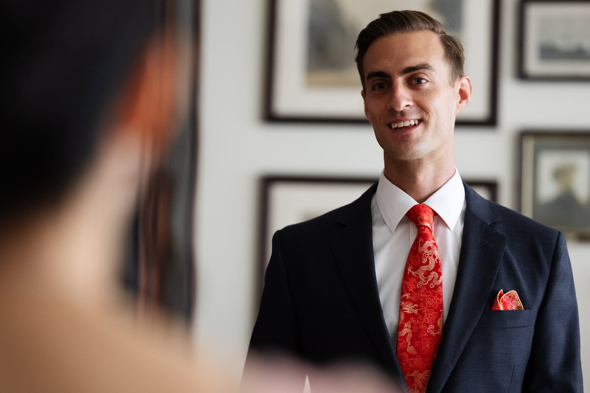 A man in a dark suit and red patterned tie stands indoors at No. 4 Hamilton Place, smiling, with framed pictures hanging on the wall behind him—a stylish setting perfect for a London wedding.