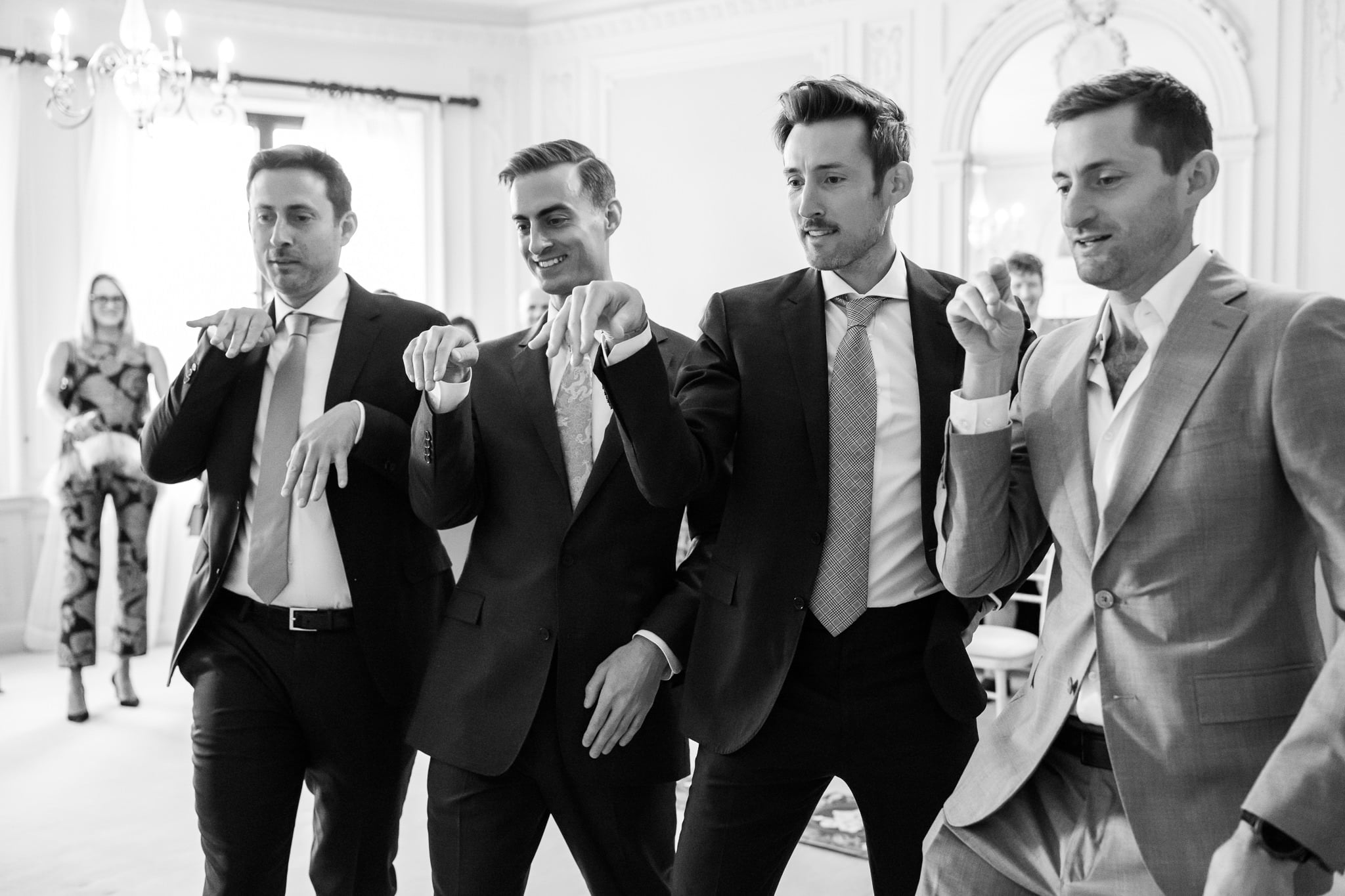 Four men in suits pose playfully with their arms bent at the wrists, imitating animal paws, in a well-lit, elegant room at No. 4 Hamilton Place. A woman stands in the background at this stylish London wedding.