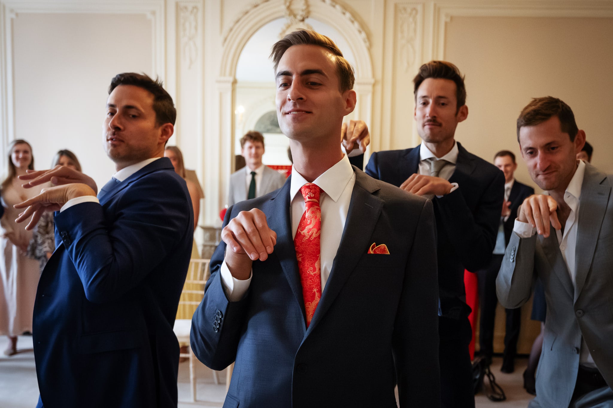 Four men in suits stand in a formal room at No. 4 Hamilton Place, holding their hands up with bent wrists and fingers together, mimicking dinosaur-like poses. People in the background watch and smile at this playful London wedding moment.