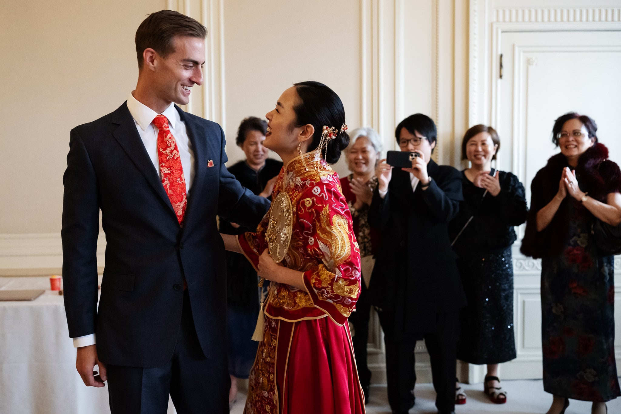 A groom in a suit and a bride in a traditional red dress smile at each other during their London wedding at No. 4 Hamilton Place, while guests in the background watch and applaud.