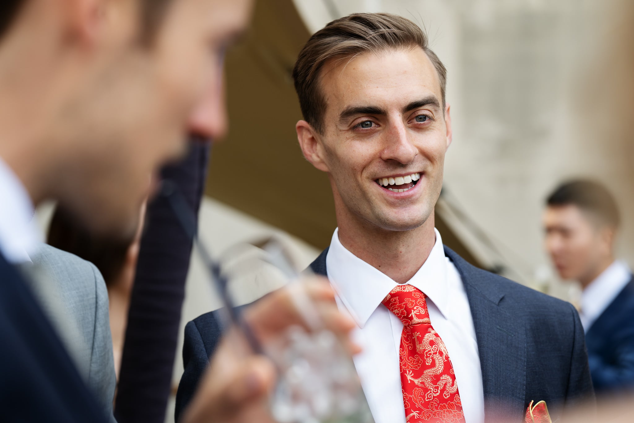 A man in a suit with a red patterned tie smiles while talking to others at an outdoor London wedding. Another person in focus is drinking from a glass with a straw at No. 4 Hamilton Place.
