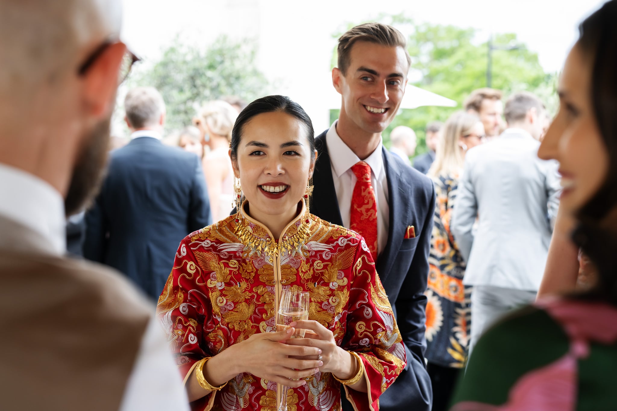 At a London wedding at No. 4 Hamilton Place, a woman in a red and gold traditional outfit holding a glass stands beside a man in a suit at a formal outdoor gathering, with other guests mingling in the background.