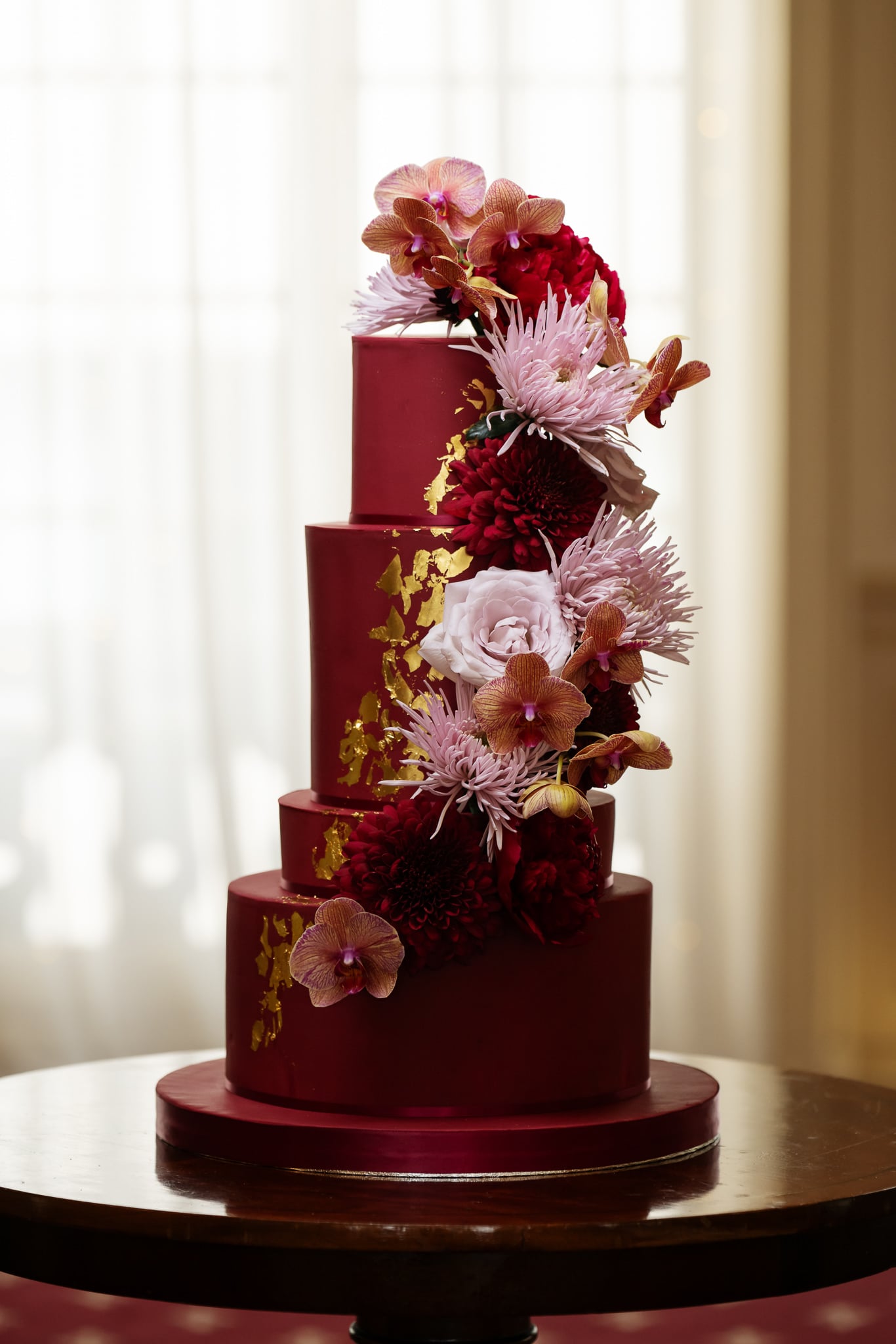 A four-tiered burgundy cake with gold leaf accents and vibrant red, pink, and white flowers, beautifully displayed on a round wooden table at No. 4 Hamilton Place for a stunning London wedding.