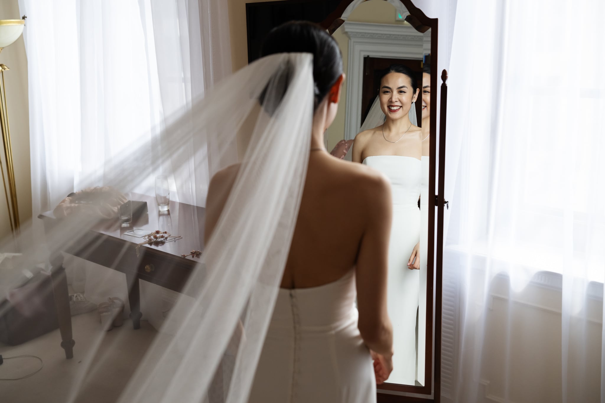 A bride in a white dress and veil stands smiling at her reflection in a well-lit room at No. 4 Hamilton Place, capturing the joy of her London wedding by the desk and window.