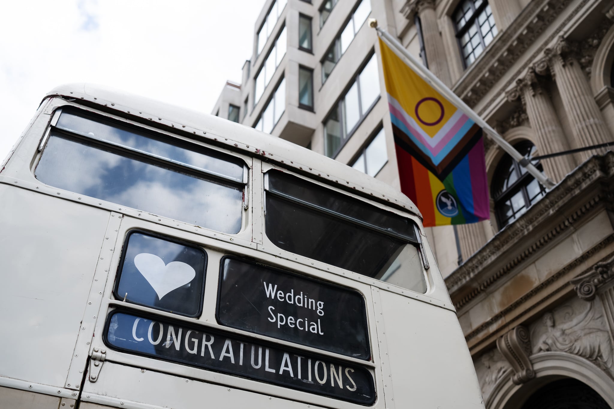 A white double-decker bus with "Wedding Special" and "Congratulations" signs is parked near No. 4 Hamilton Place, adding charm to this London wedding scene beside a building with a Progress Pride flag.