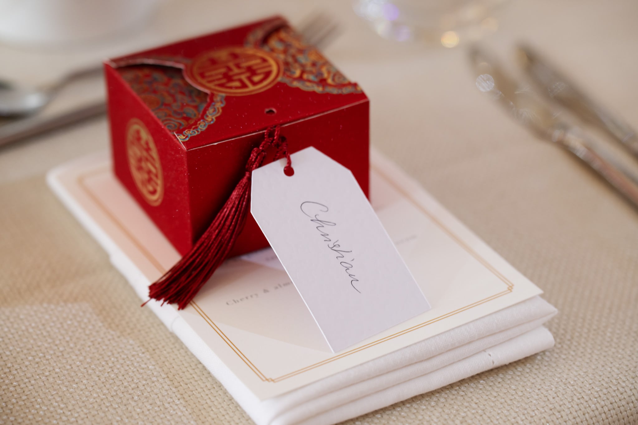 A red decorative box with a tassel and a name tag reading "Christian" sits on a folded napkin atop a cream-colored tablecloth, ready to delight guests at a stylish London wedding at No. 4 Hamilton Place.
