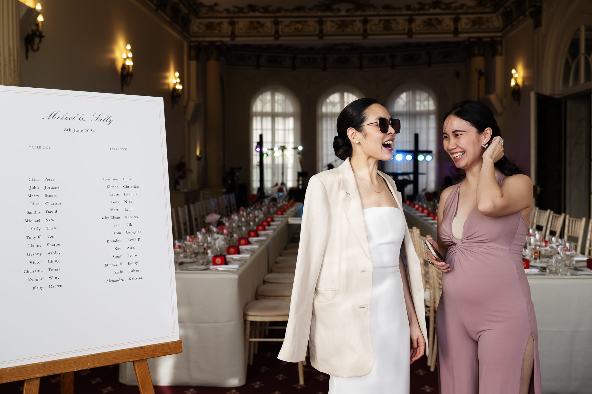 Two women stand and smile next to a large wedding seating chart in an elegant dining hall with high ceilings and chandeliers at No. 4 Hamilton Place, a stunning venue for a classic London wedding.