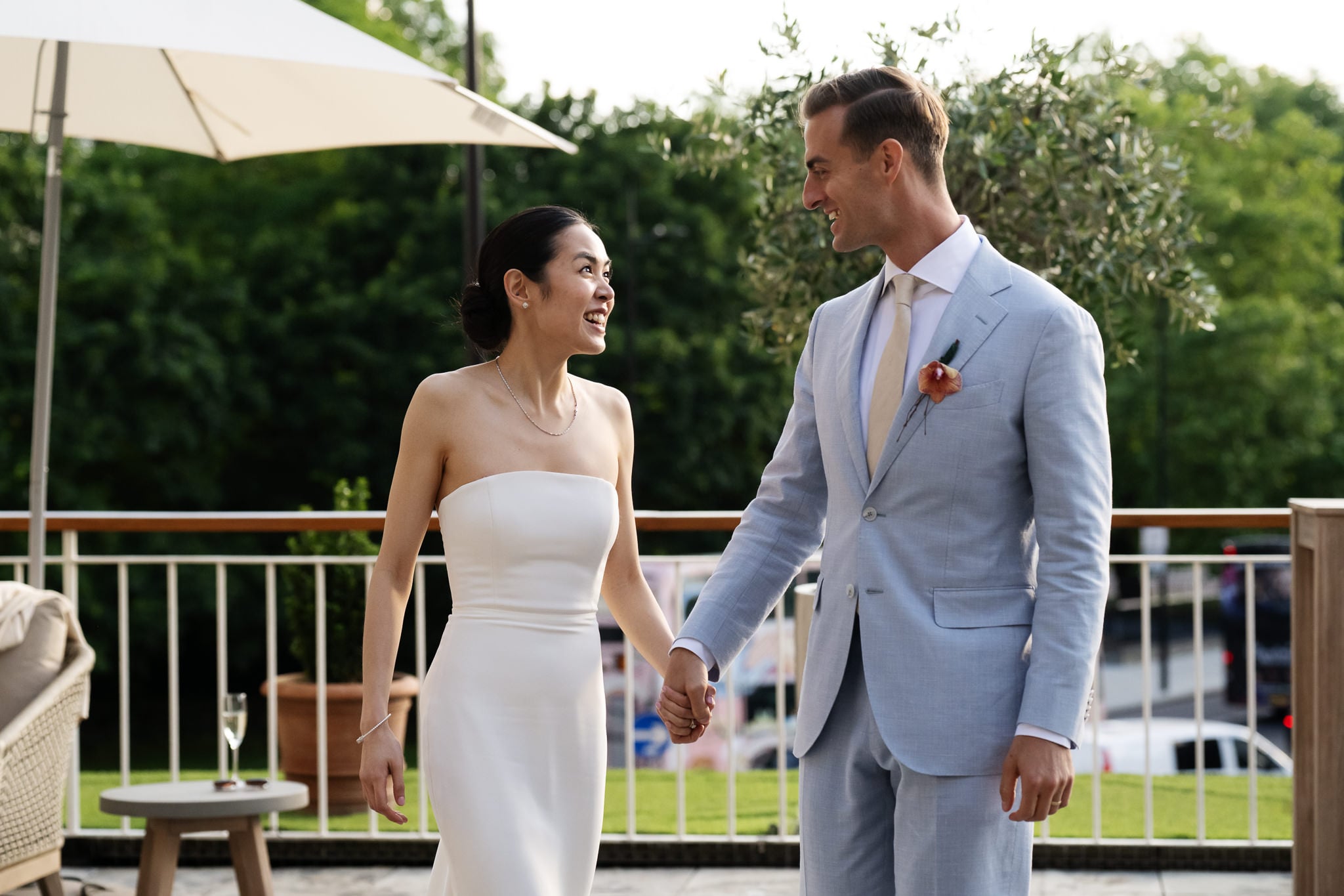 A couple dressed in formal attire, holding hands and smiling at each other outdoors amid greenery and an umbrella—capturing the charm of a London wedding at No. 4 Hamilton Place.