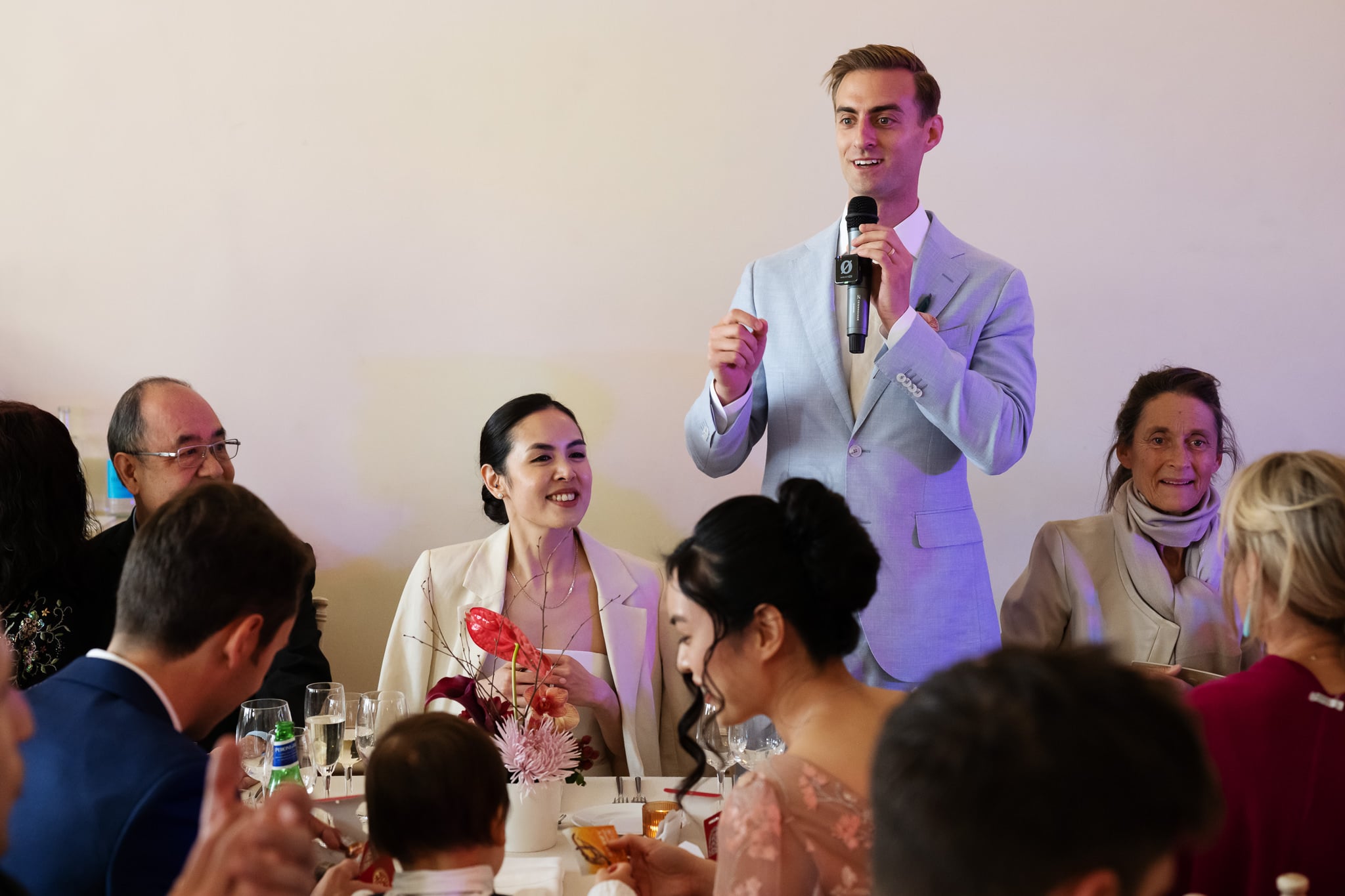 At No. 4 Hamilton Place, a man in a light suit speaks into a microphone at a banquet table with several guests, including a woman in white beside him, during an elegant London wedding gathering.