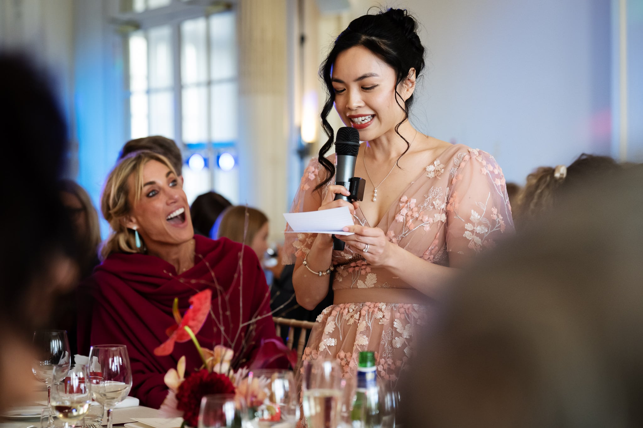 At No. 4 Hamilton Place, a woman in a pink floral dress speaks into a microphone, holding a note card, while guests seated at tables smile and listen during this elegant London wedding.