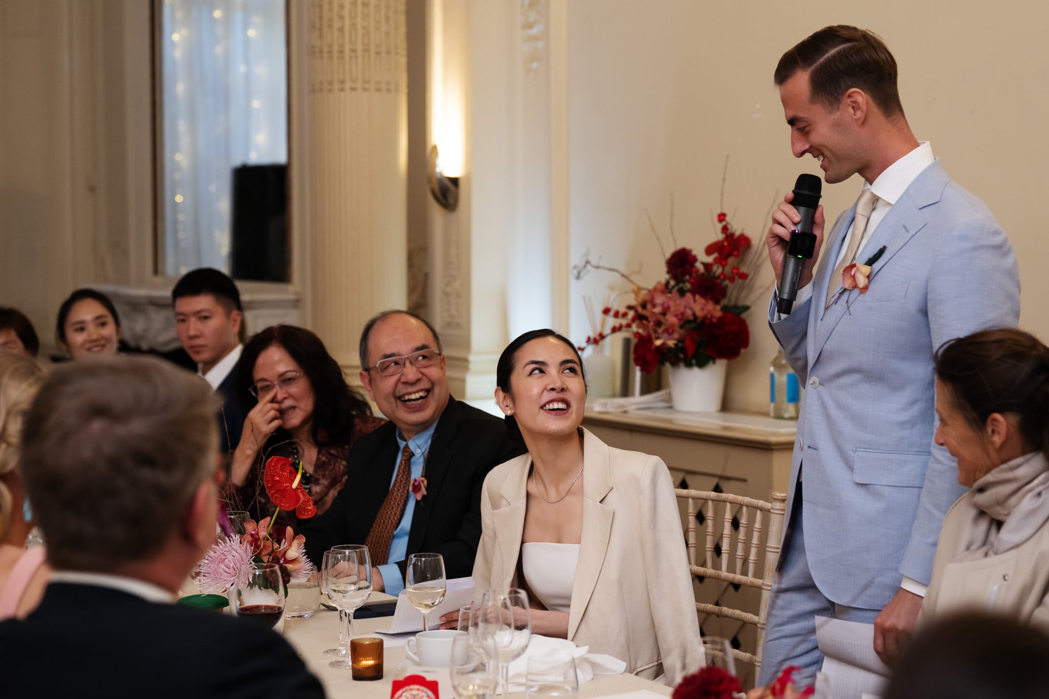 At a London wedding at No. 4 Hamilton Place, a man in a light blue suit gives a speech with a microphone at a formal event while guests seated at a table listen and smile.