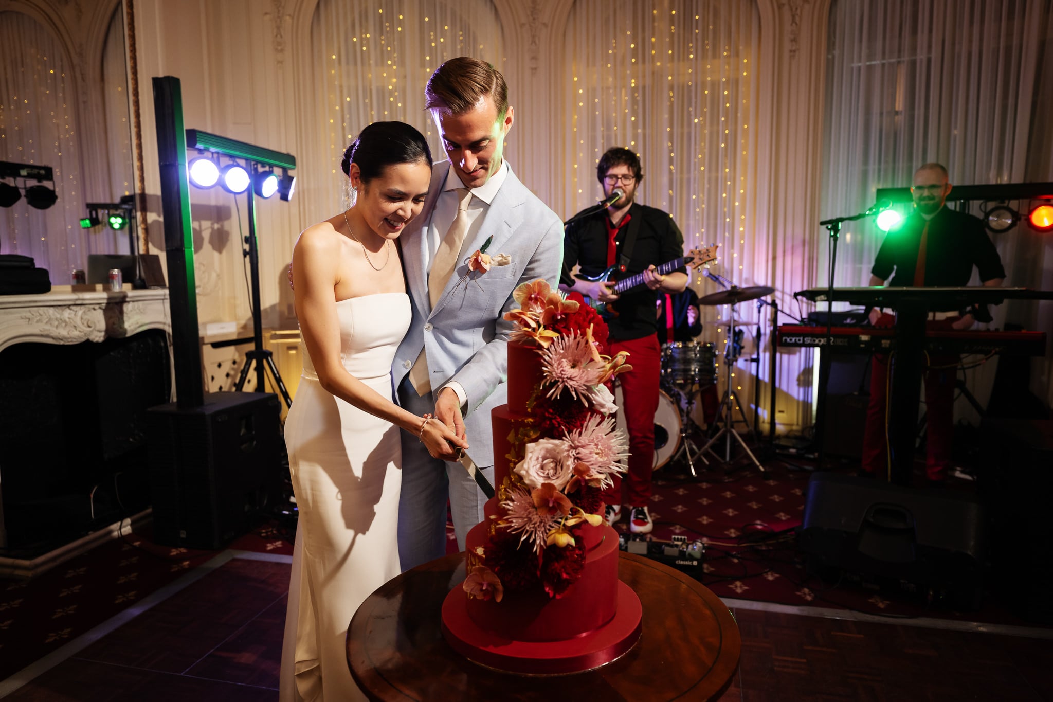 A bride and groom cut a red wedding cake with floral decorations at their London wedding, while a live band performs in the background at No. 4 Hamilton Place’s beautifully decorated indoor venue.