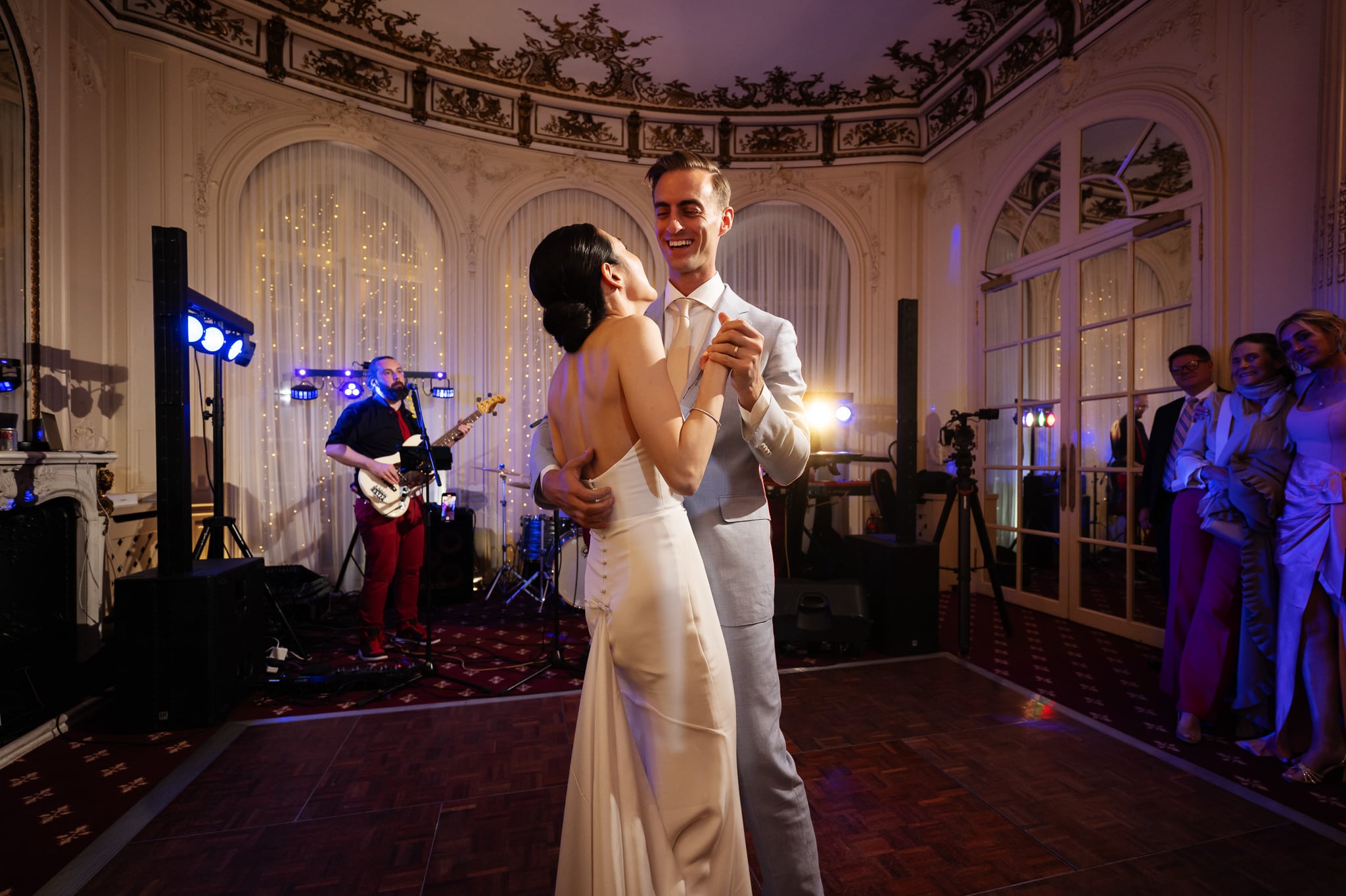 A bride and groom share their first dance in the grand, ornate ballroom of No. 4 Hamilton Place at a romantic London wedding, as a live band performs and guests watch in the background.