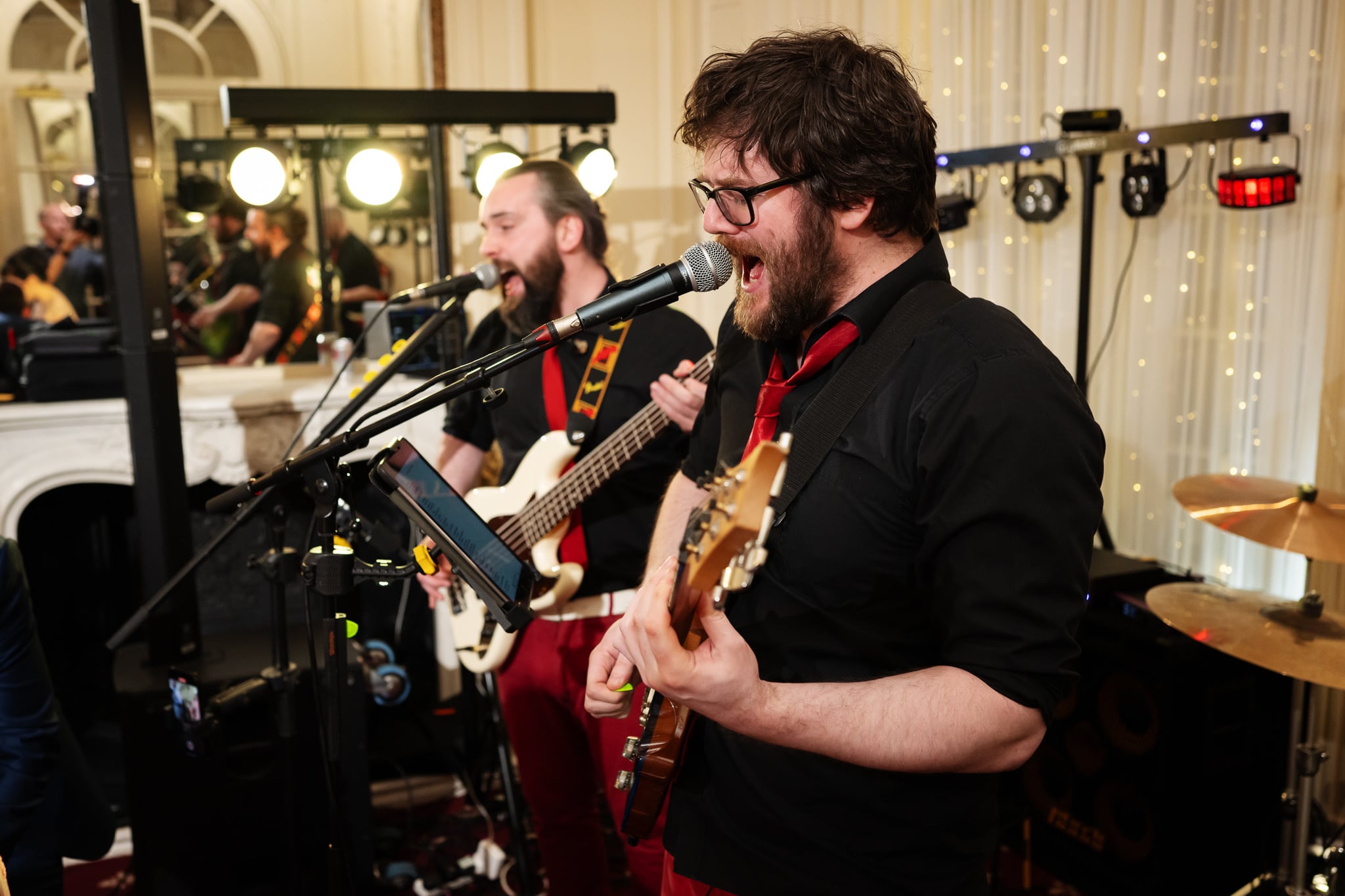 Two musicians perform indoors at No. 4 Hamilton Place, singing into microphones while playing electric guitars. The background features musical equipment and decorative lights, setting a stylish scene for a London wedding.